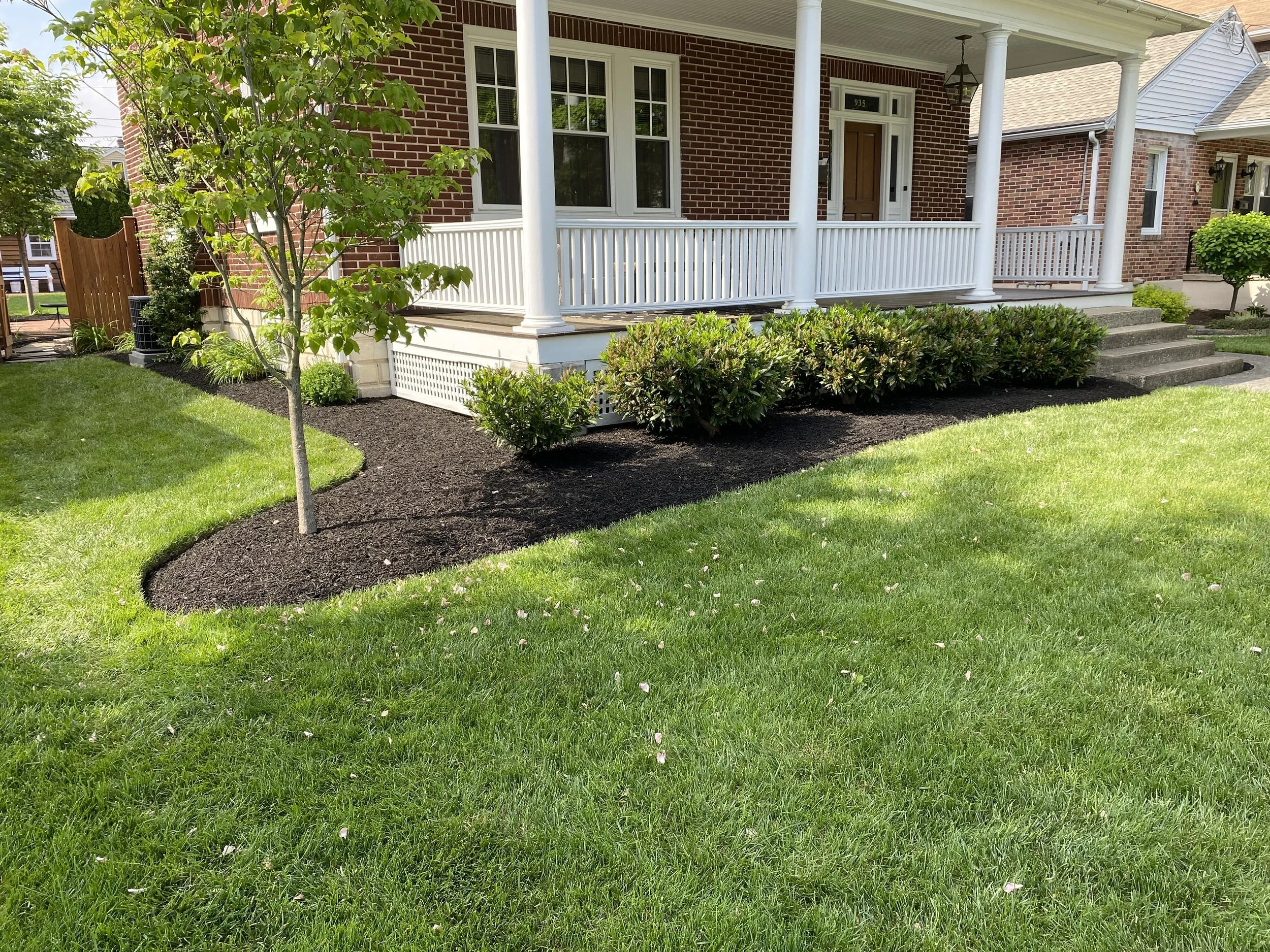 Front yard of a brick house with a porch, steps, and a white railing. There are trees, bushes, and a lawn in the yard.