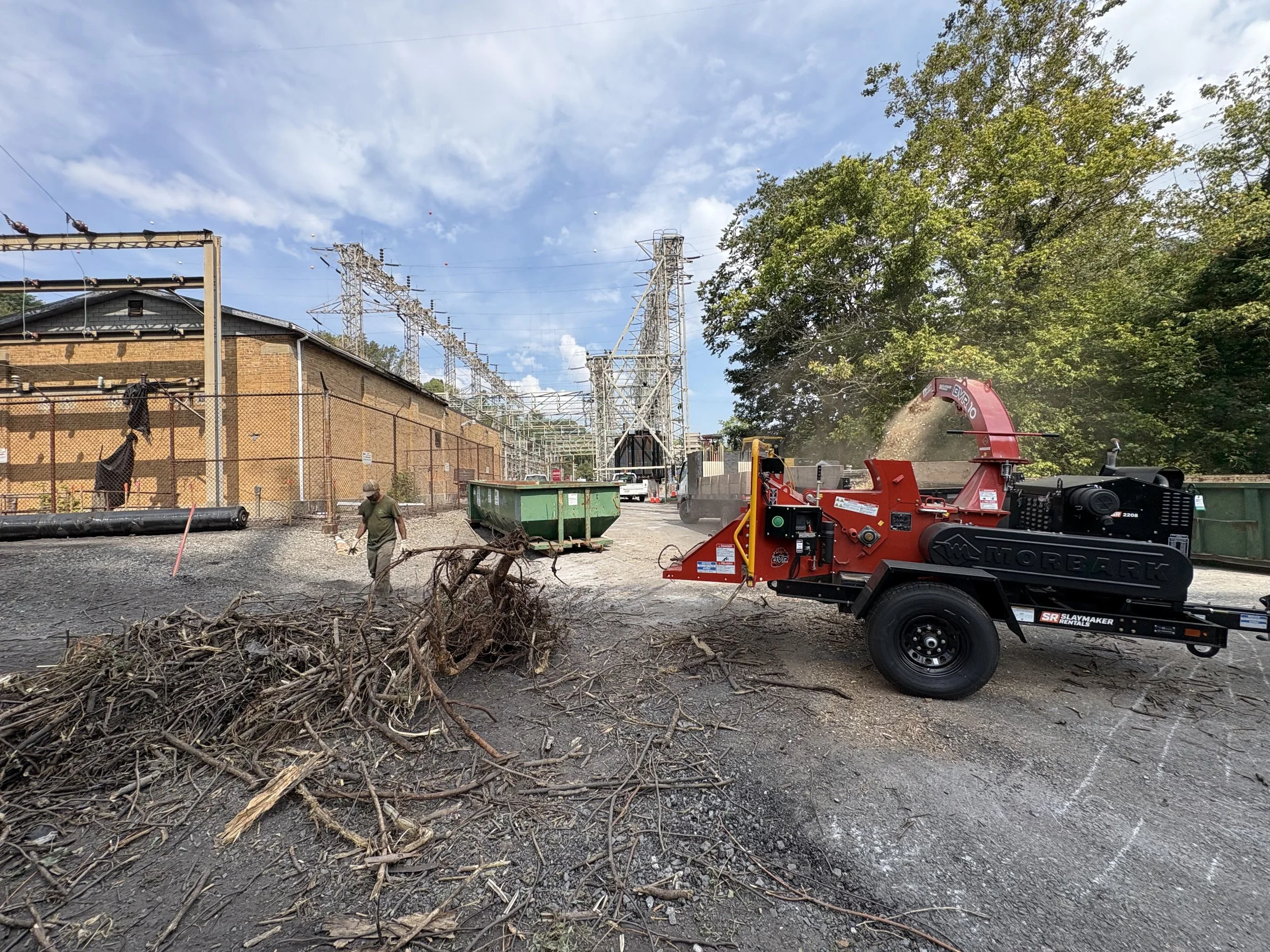 Construction site with workers, machinery, trees, and power lines under a cloudy sky.