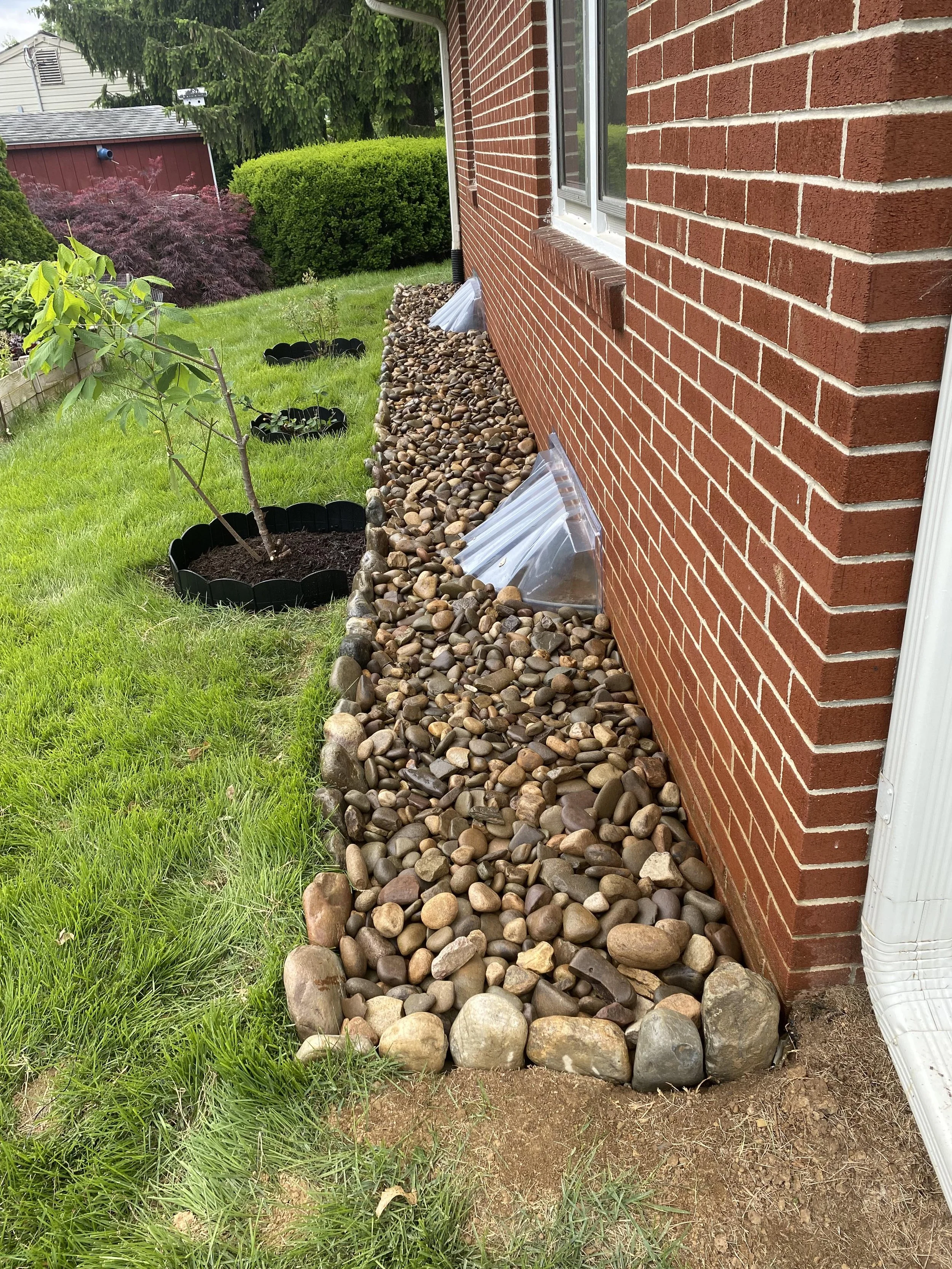 Side of a brick house with a dug-up garden bed filled with rocks and stones, some plastic sheeting, and a small tree in the yard with green grass and bushes nearby.