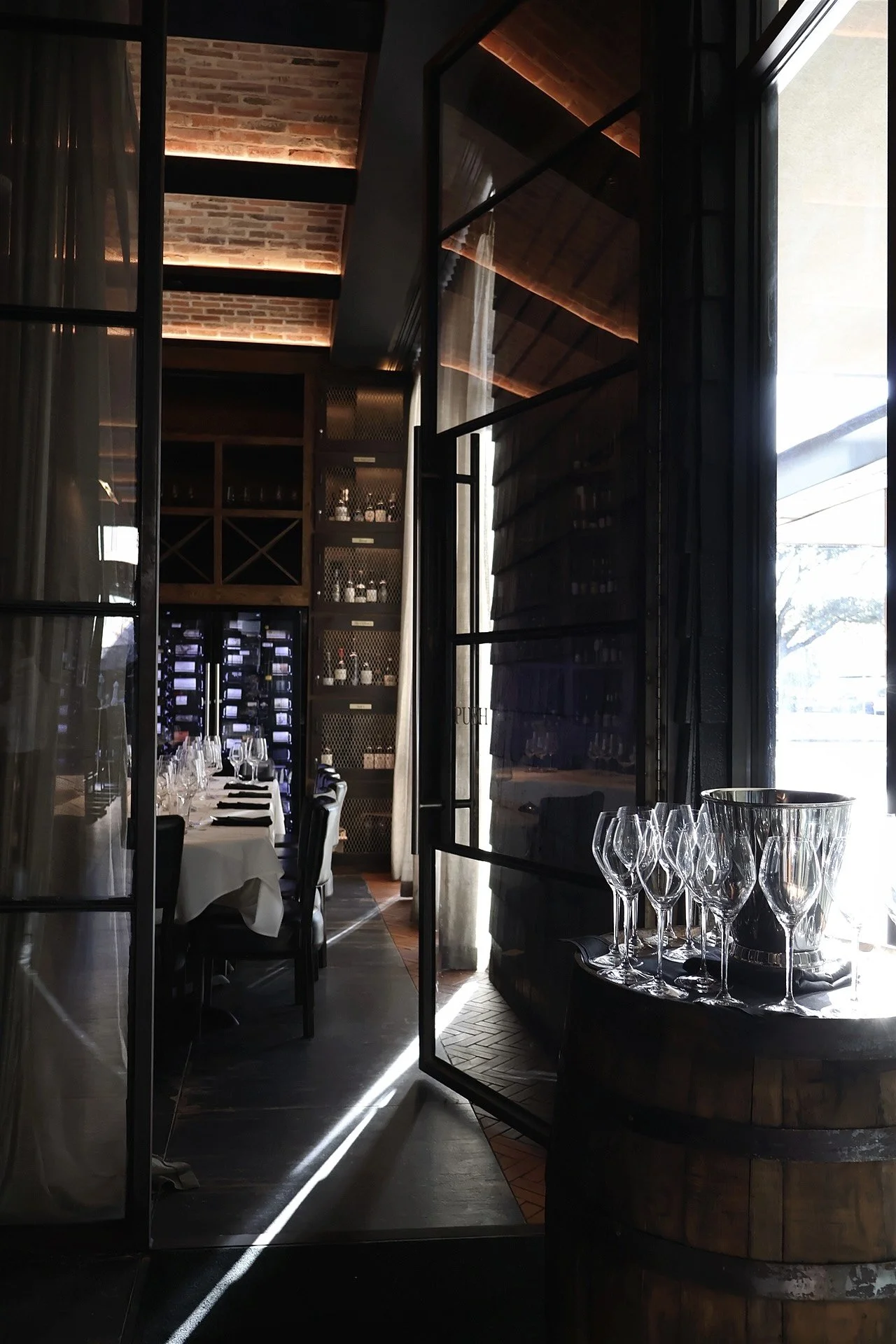 Elegant restaurant interior with a table set for dining, wine glasses on a barrel, and a wine storage area with bottles behind glass doors. Large windows allow natural light to illuminate the space.