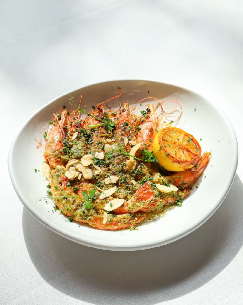 Shrimp dish garnished with herbs, sliced almonds, and accompanied by a grilled lemon half in a white bowl.