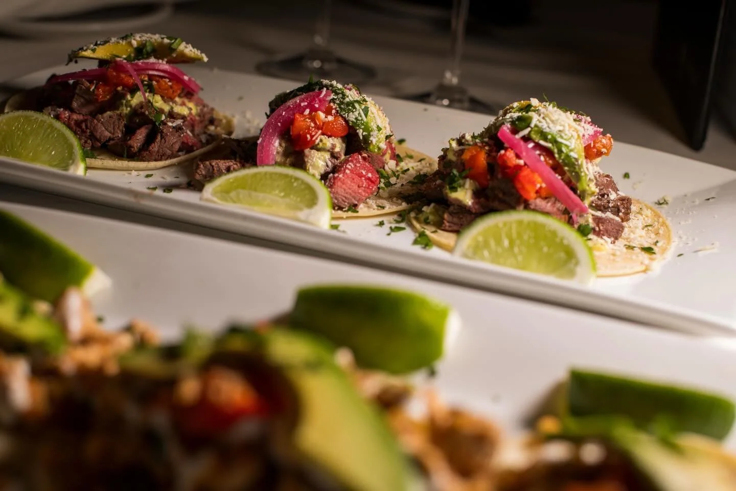 Three authentic Mexican tacos with various meats, vegetables, and toppings, garnished with lime wedges on a white rectangular plate.
