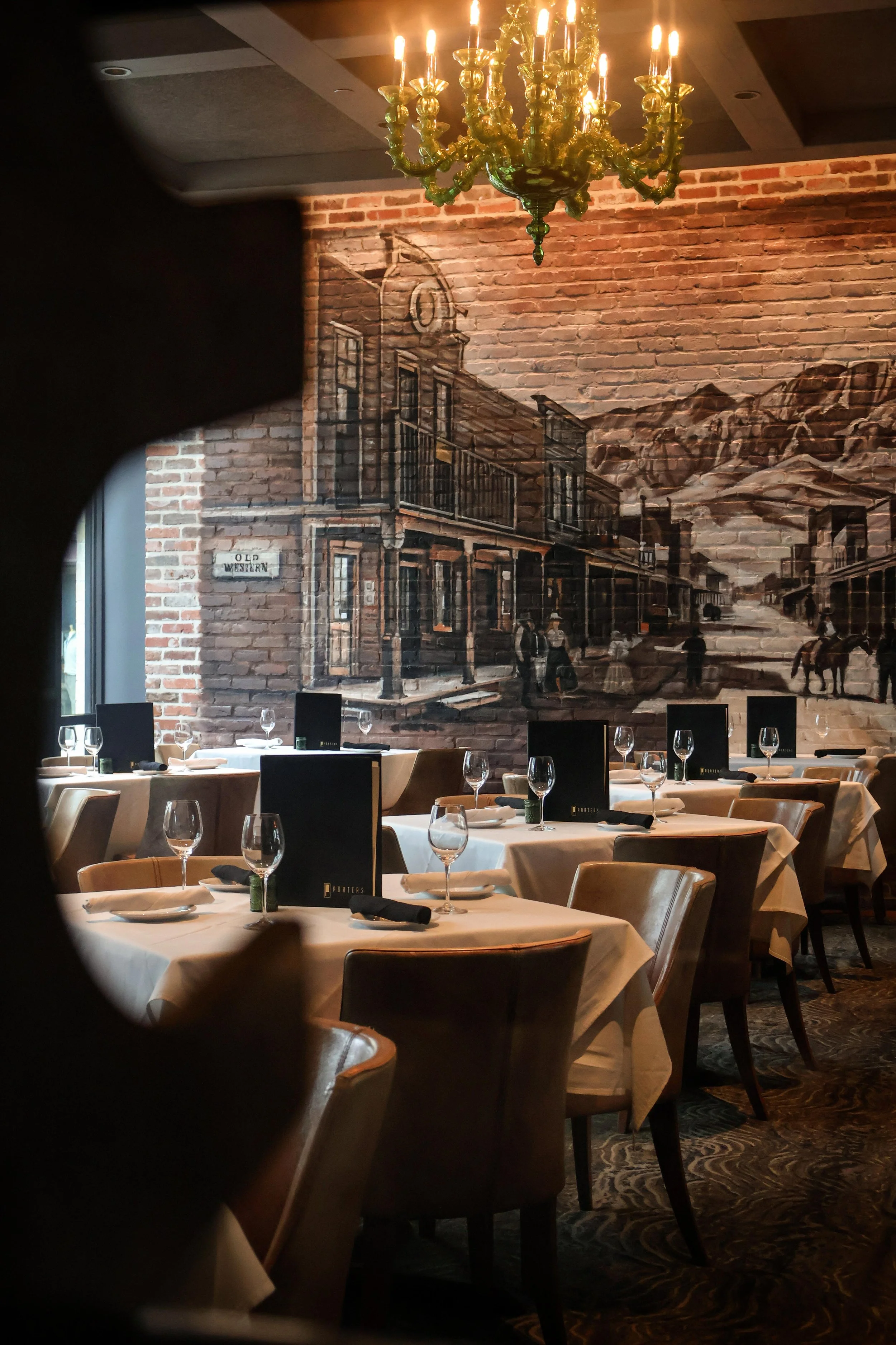 A dimly lit restaurant with several round tables set with white tablecloths, wine glasses, and black menus. A large brick wall features a mural of a Western town scene, and a green chandelier hangs from the ceiling. The photo is taken through a circular frame or cutout.