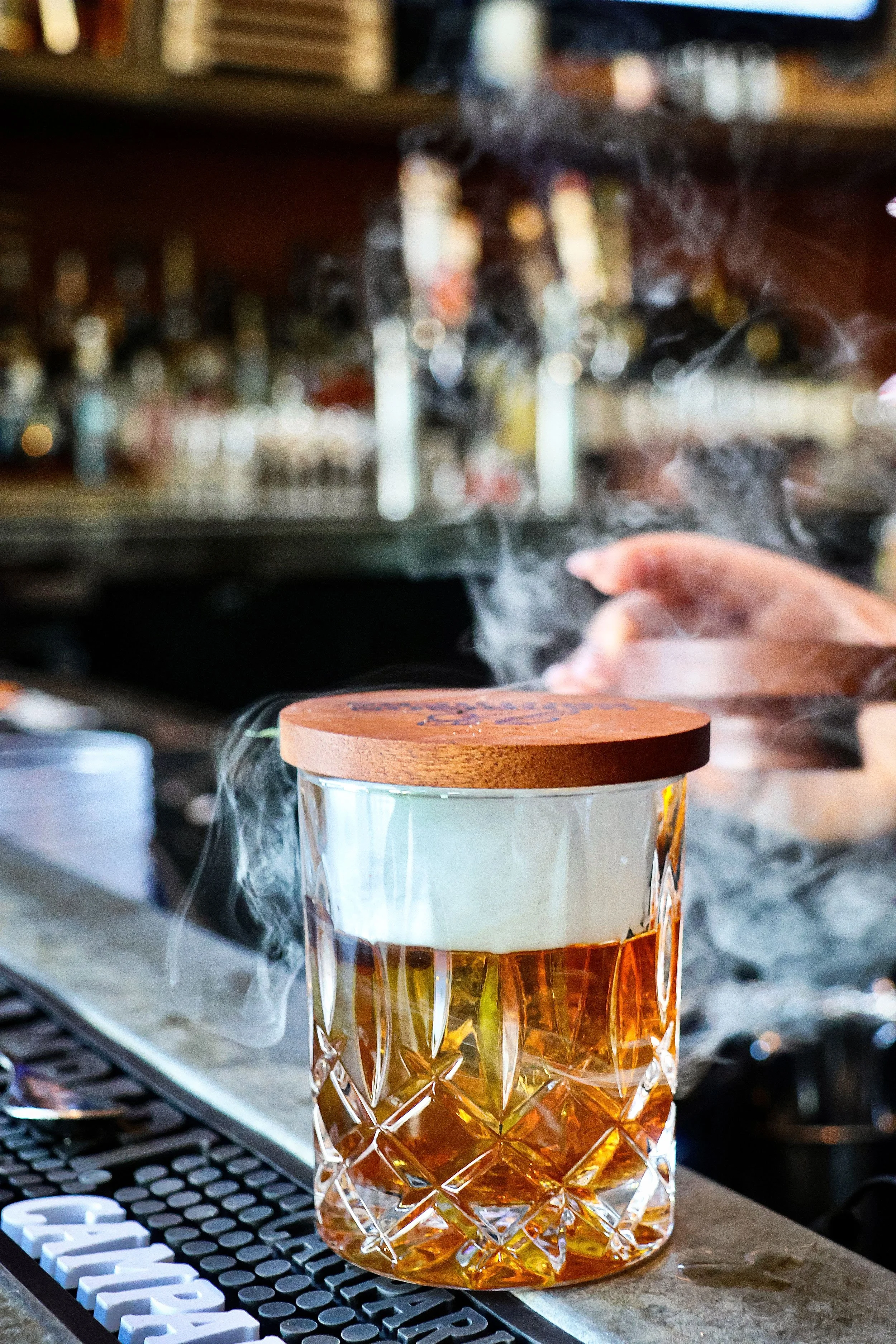 A glass of whiskey with a diamond pattern on the glass, a white label, and a wooden lid, sitting on a surface near a keyboard, with steam or smoke in the background.