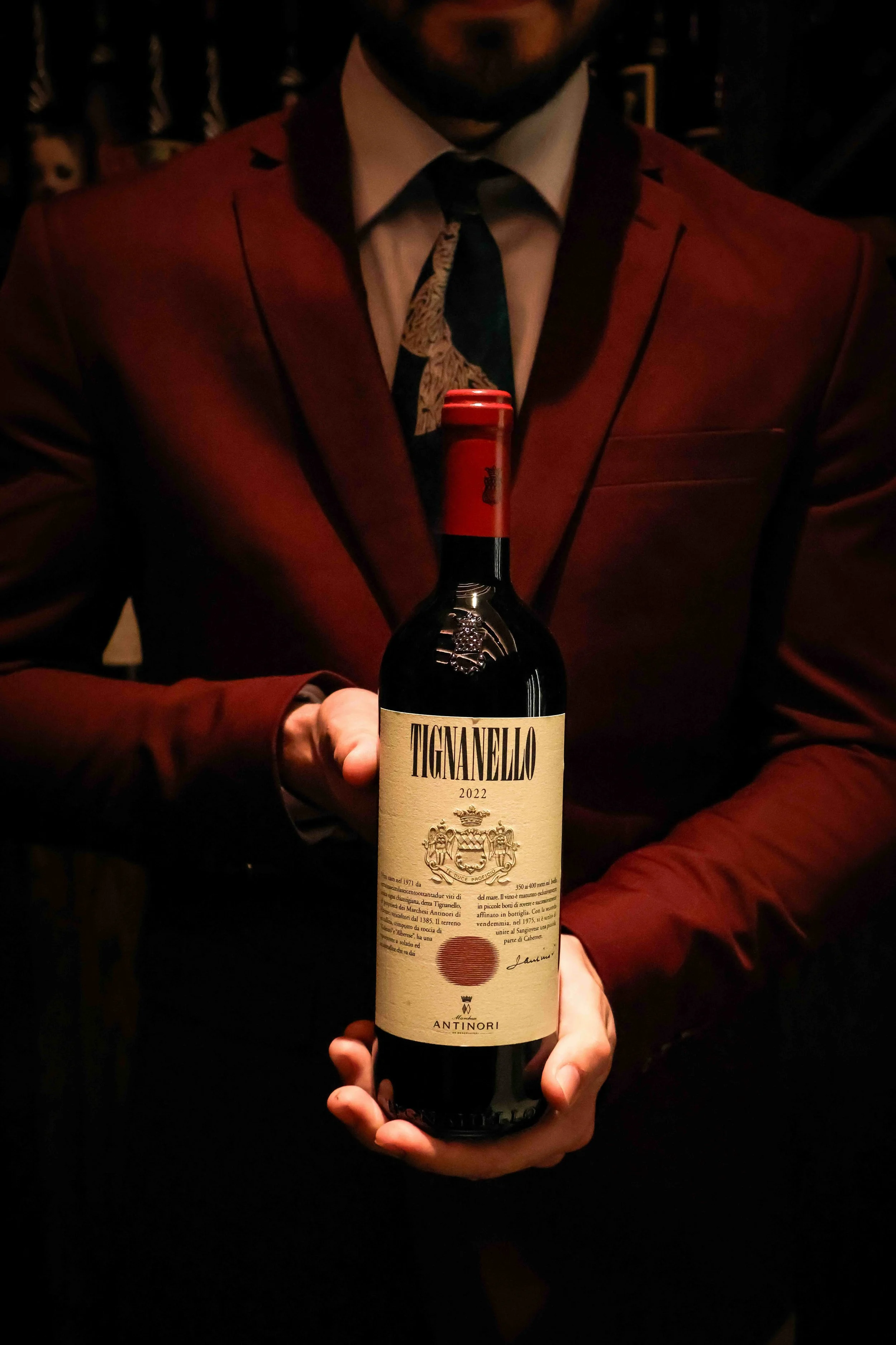 A person dressed in a dark red suit holding a bottle of wine with a beige label and red cap. The person's face is partially visible, and they are wearing a dress shirt and tie with a patterned design.