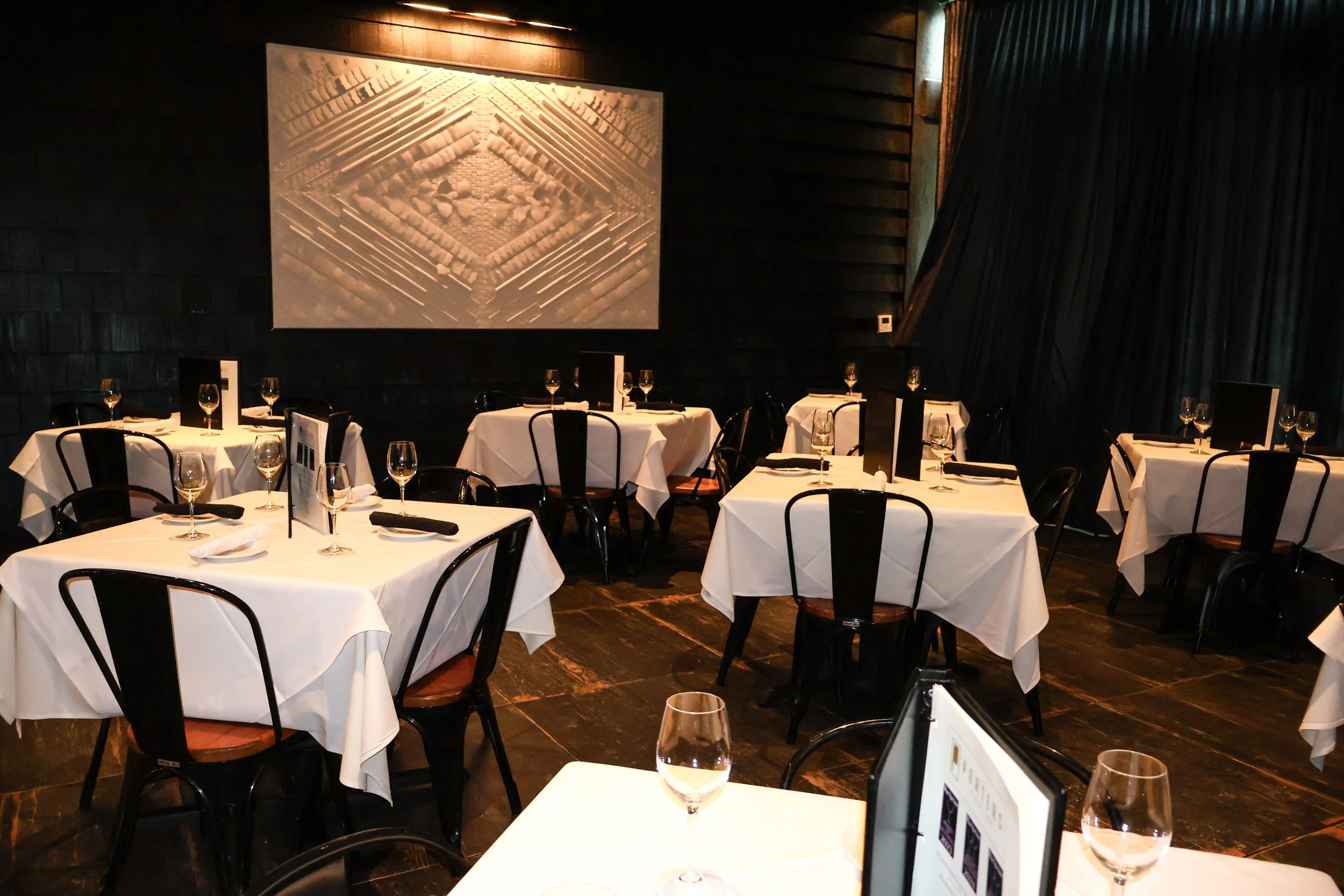 Privately set restaurant with round tables covered with white tablecloths, each set with wine glasses, napkins, and menus, and dark chairs surrounding them. A large abstract art installation or textured panel decorates a dark wall behind the tables, with dim lighting creating a cozy atmosphere.