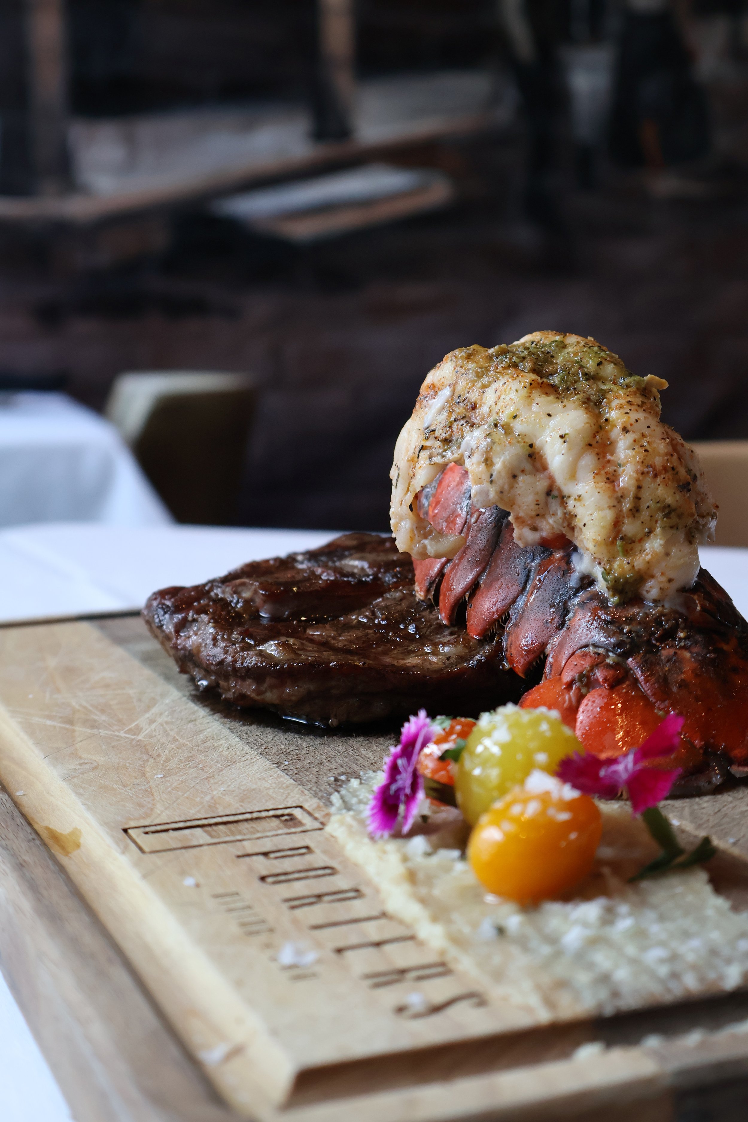 Close-up of a wooden board with a cooked steak topped with lobster tail and herb butter, garnished with colorful edible flowers and small yellow berries.