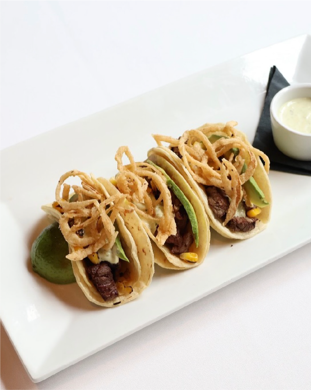 Three tacos with beef, avocado, corn, and crispy fried onions on a white rectangular plate, served with a lime wedge and a small bowl of sauce.