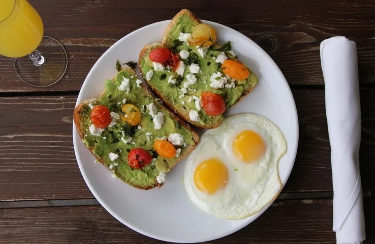 A white plate with two sunny-side-up eggs and toast topped with avocado, cherry tomatoes, and crumbled cheese.