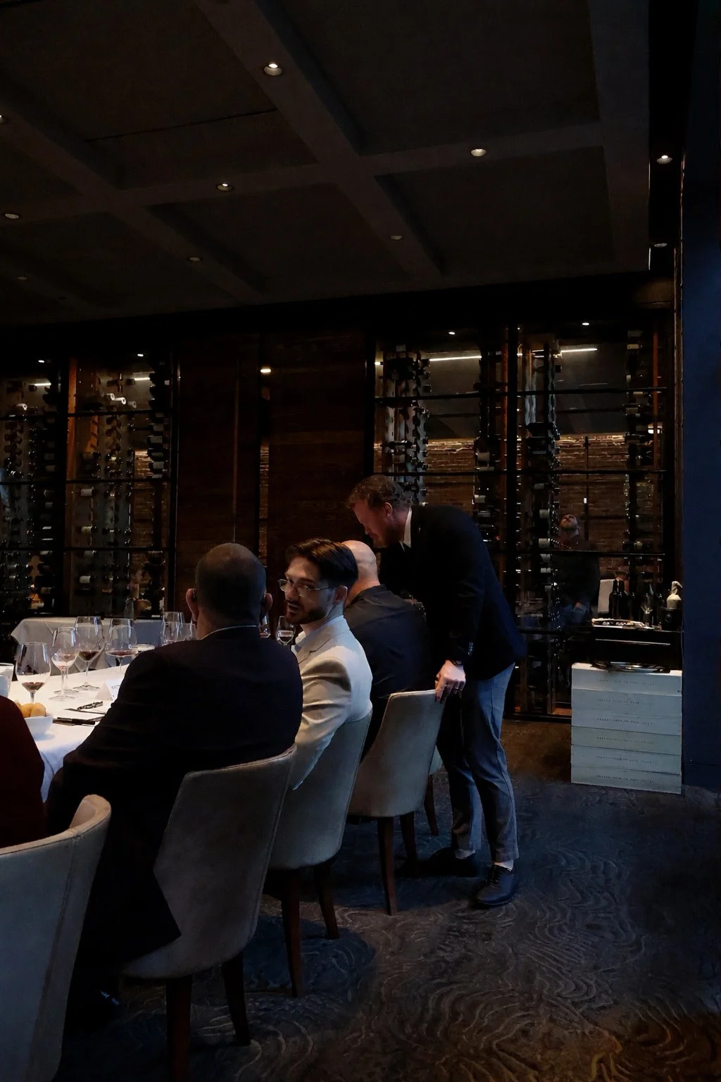People dining in a dimly lit restaurant with wine glasses on the table and a wine rack in the background.