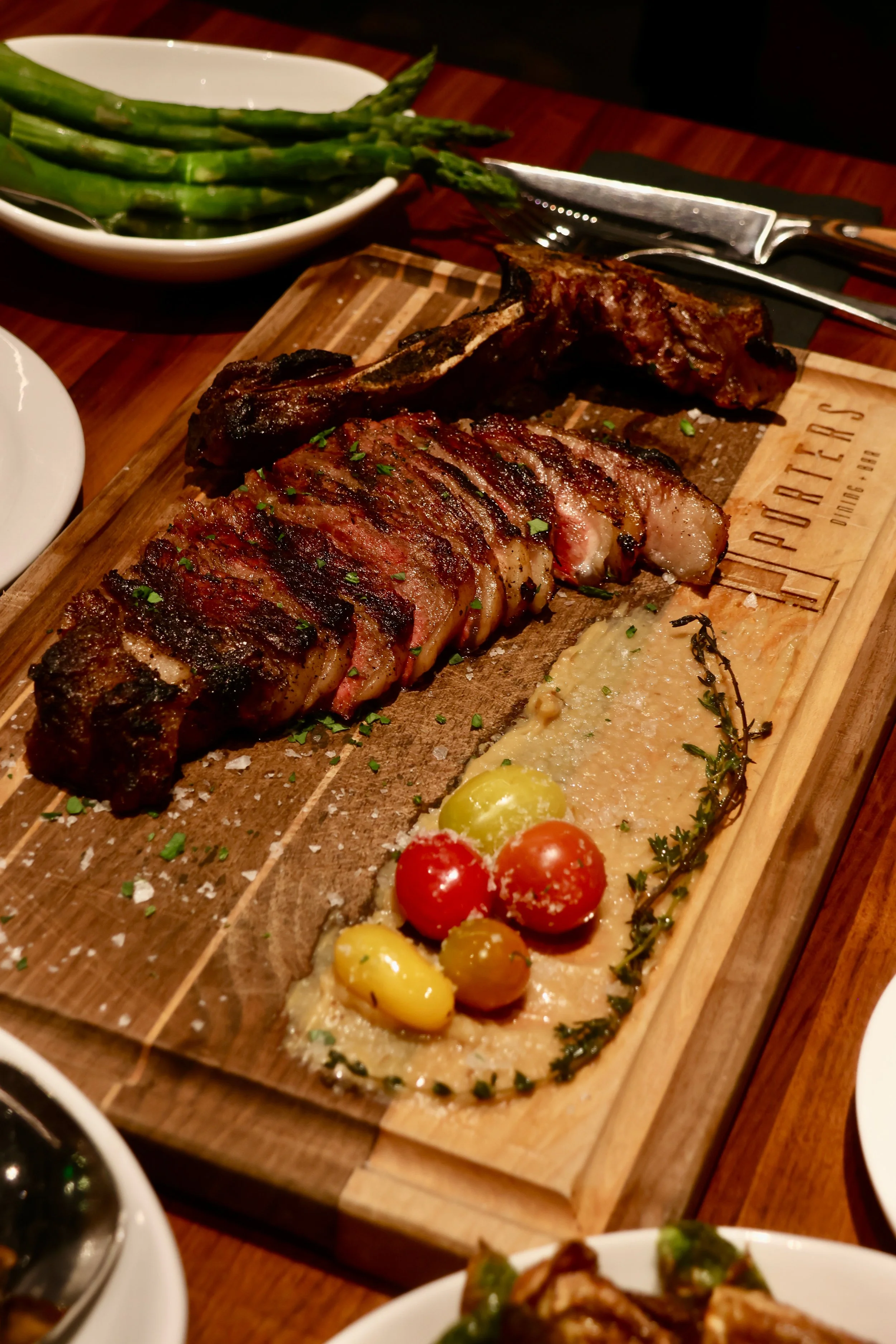 A wooden platter with grilled meat, mashed potatoes garnished with herbs, cherry tomatoes, and olives, with a side of asparagus on a white dish in the background.