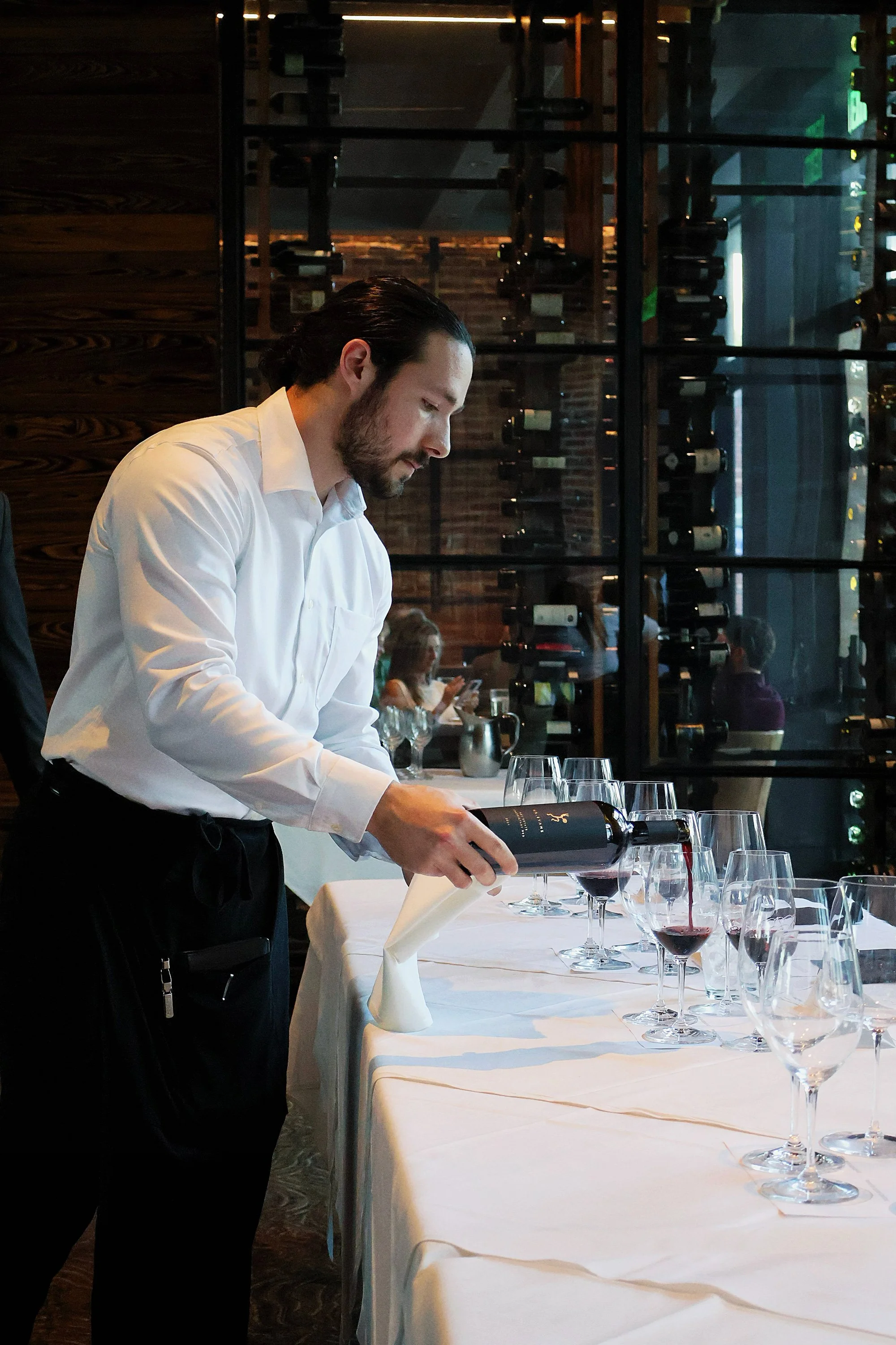 A male sommelier with dark hair and a beard pours red wine into glasses on a white tablecloth in a restaurant. The background features a wine rack and patrons seated at other tables.