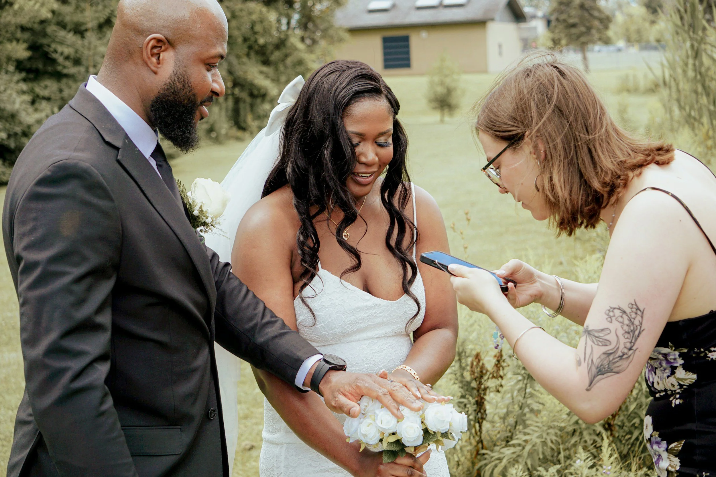 A wedding ceremony outdoors with a bride, groom, and officiant. The bride holds a bouquet of white roses, and they are exchanging vows or rings while the officiant looks on, in a green park setting.