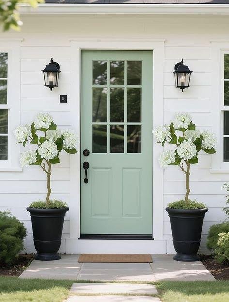 Front door painted in mint green with two potted white hydrangea plants on either side, black wall lanterns above, white siding, and a small welcome mat.