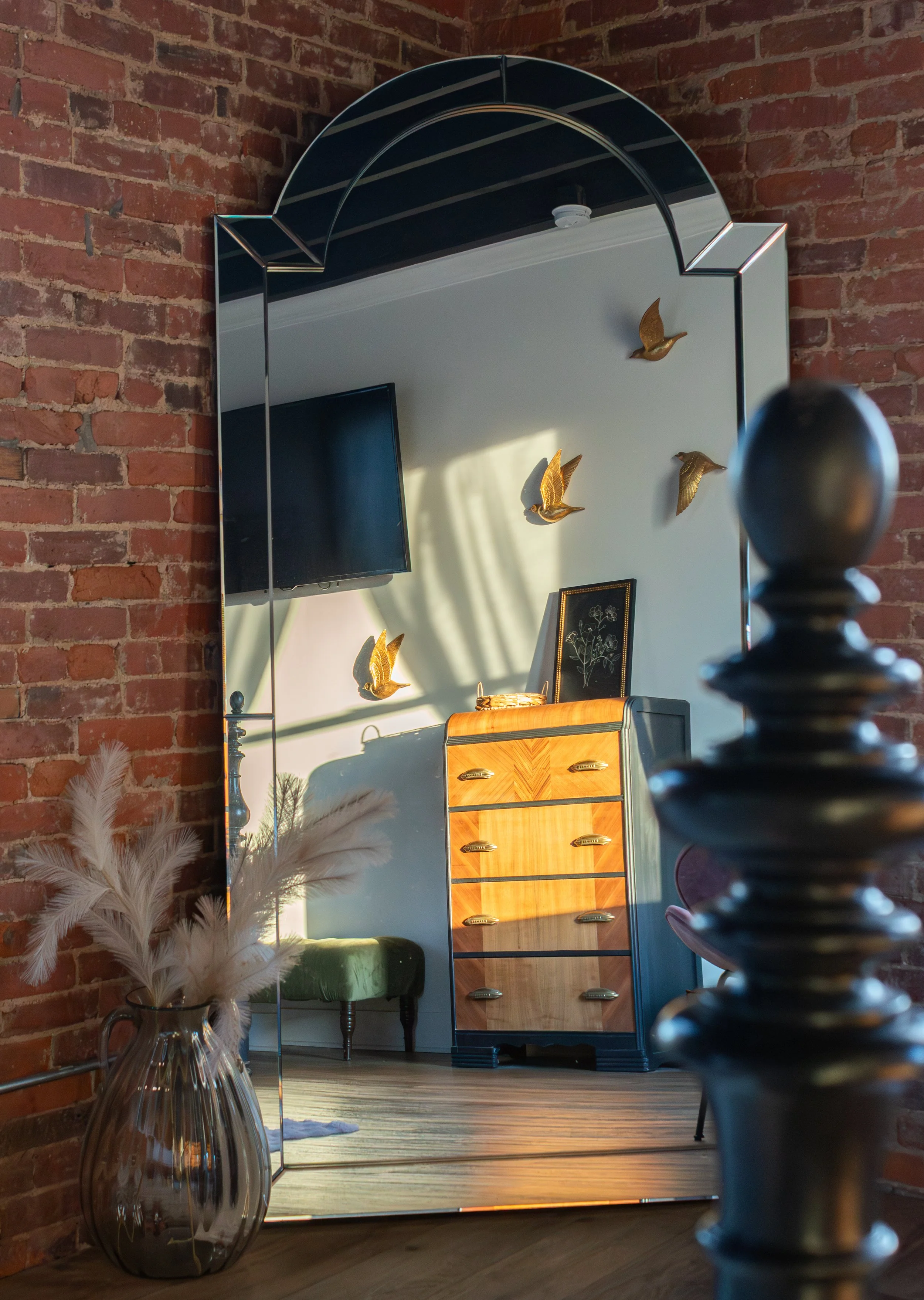 A large mirror with a curved top reflecting a brick wall, a wooden dresser, wall-mounted golden bird sculptures, and a television in a room with wooden flooring and decorative items including a glass vase with white feathers.