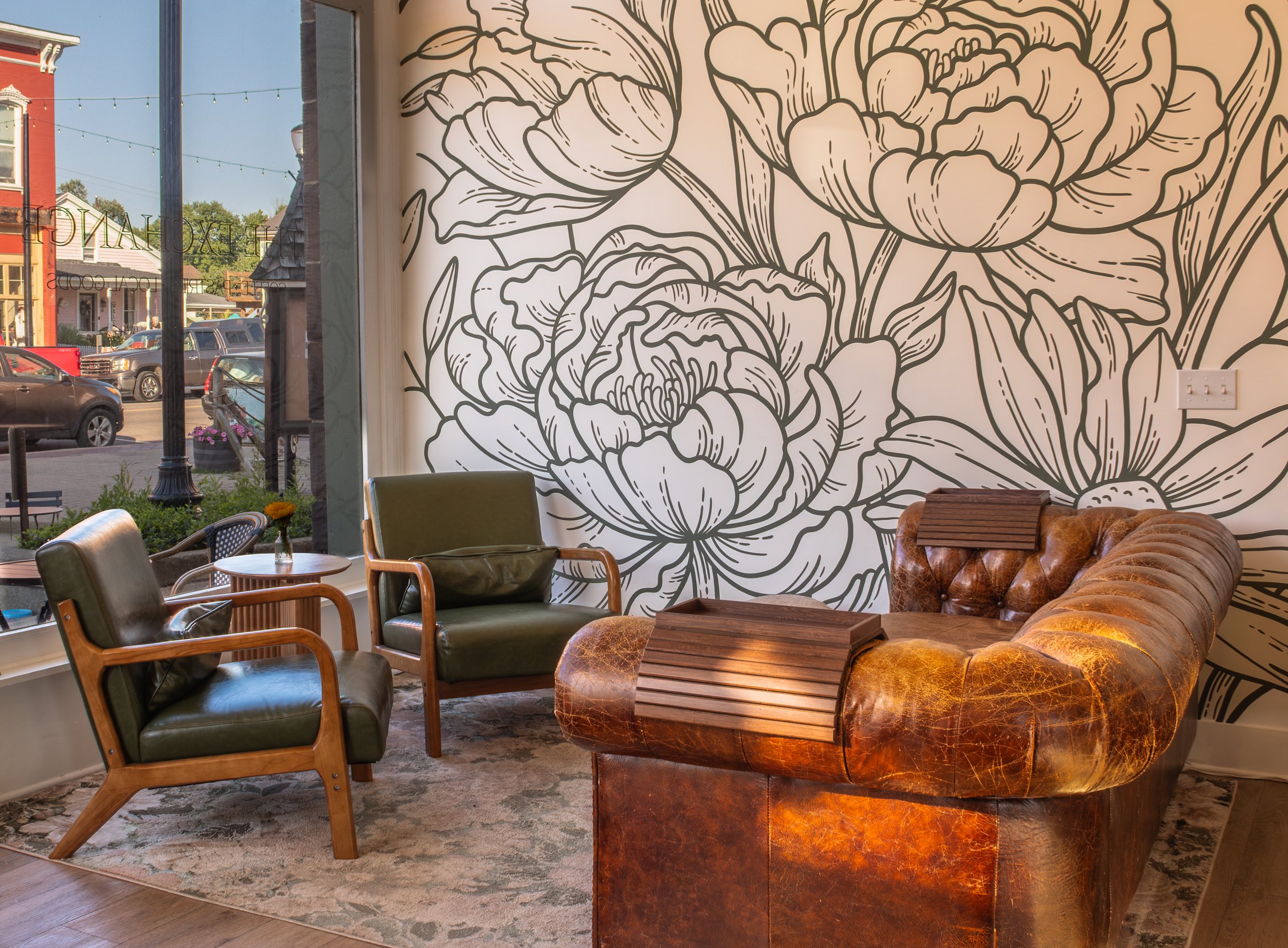 Interior of a cozy cafe with a large black and white floral mural on the wall, vintage leather and wooden chairs, a small round table with a vase, and large windows showing a street view outside.