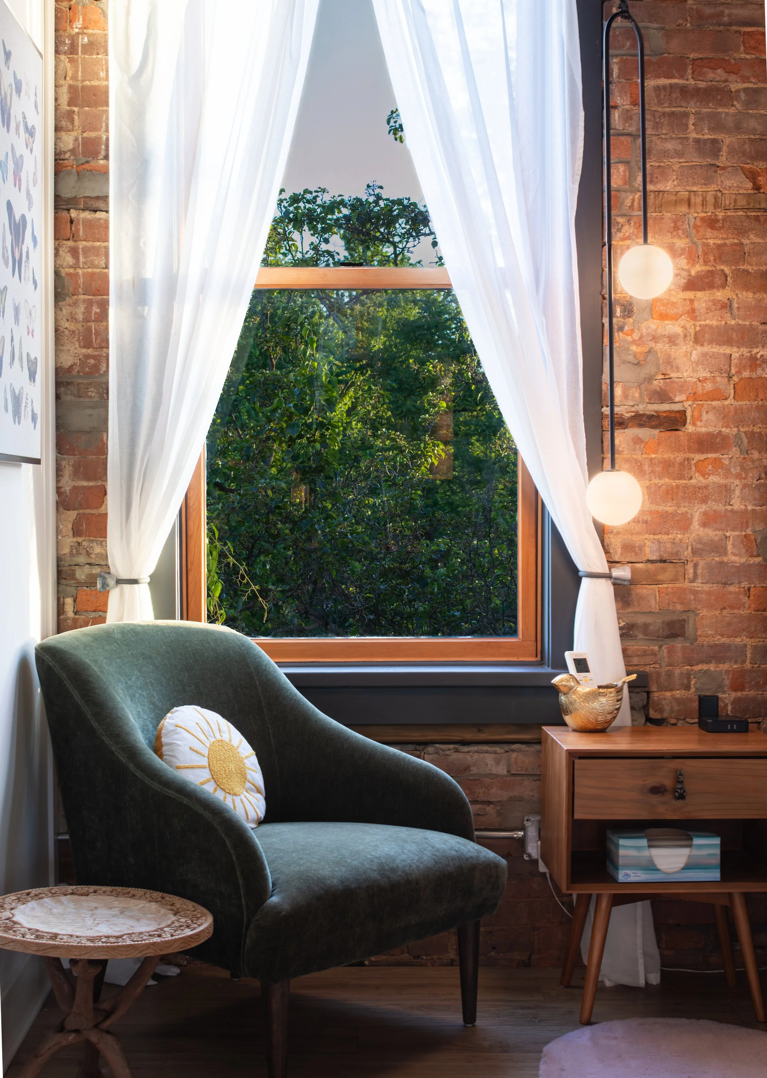 A cozy living room corner with a green velvet armchair that has a round pillow with a sun design, a small wooden side table with a decorative bowl and a tissue box, a window with white curtains showing green trees outside, and a brick wall with hangi