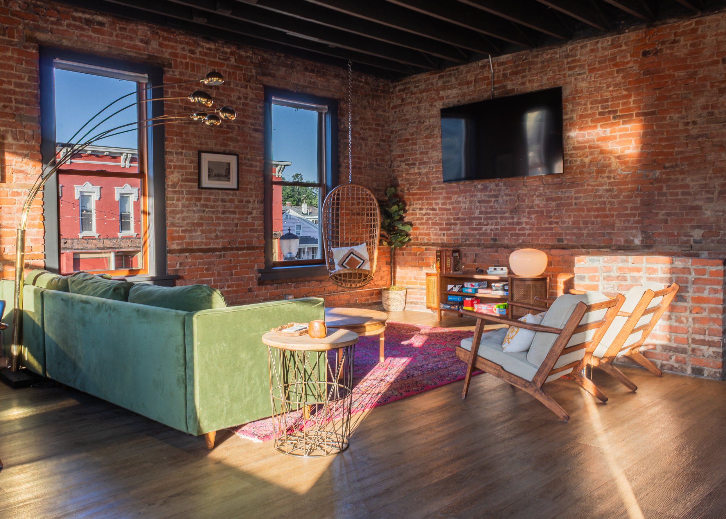 Living room with exposed brick walls, large windows, a green velvet sofa, a wicker hanging chair, a pink patterned rug, a wooden side table with a lamp, a bookshelf, and a flat-screen TV mounted on the wall.
