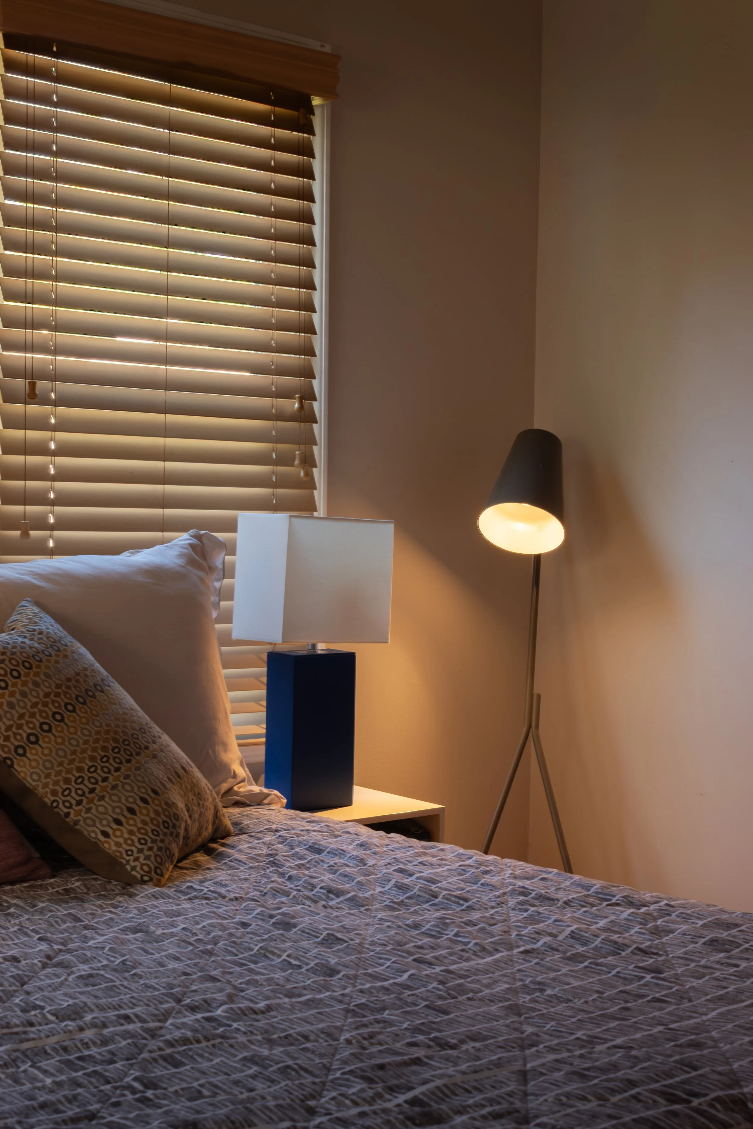 A bedroom with a bed, pillows, a nightstand with a lamp, and a floor lamp, with closed window blinds letting in some light.