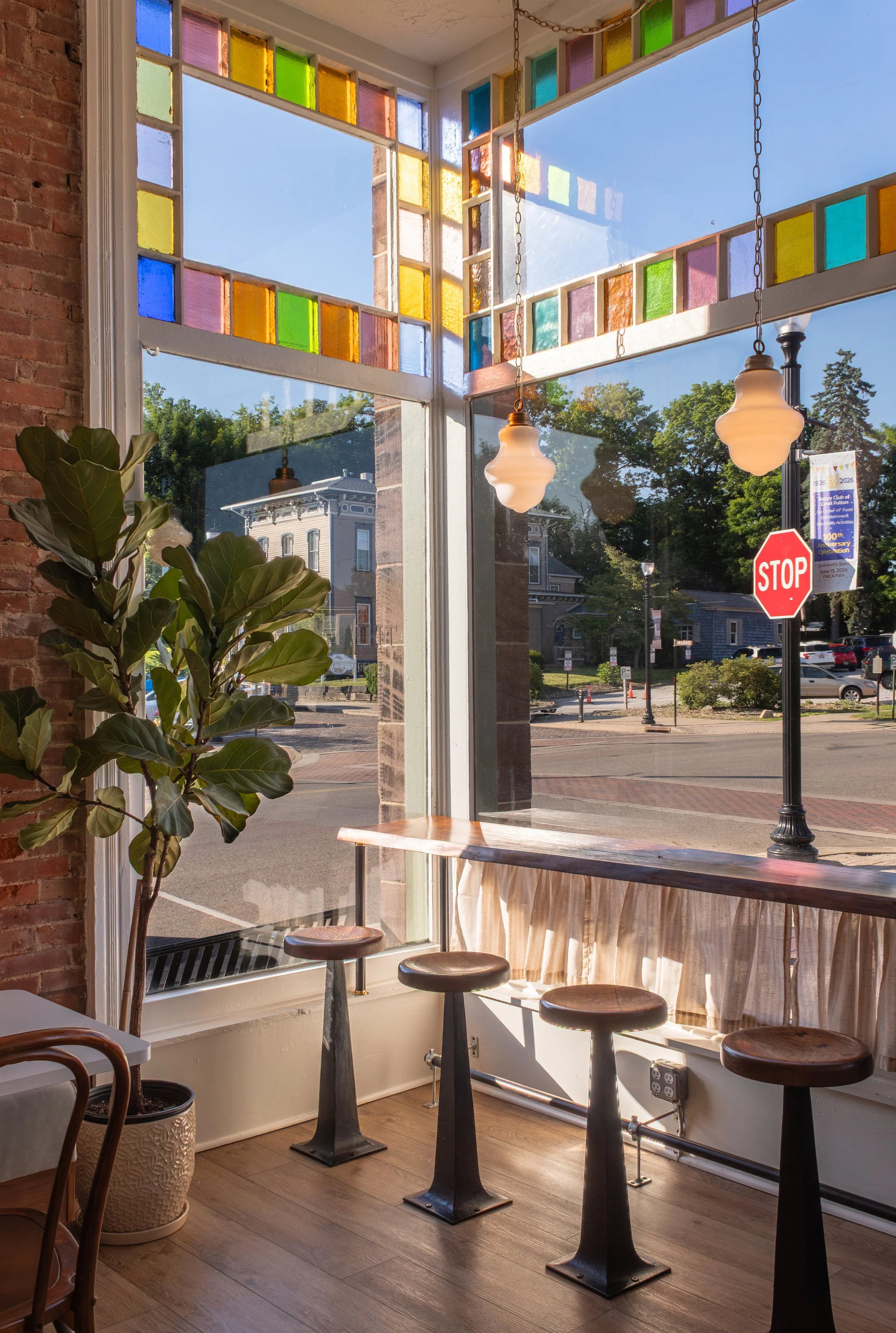 Interior of a cozy cafe with a large window featuring colorful stained glass accents. Outside, a street scene with historic buildings, trees, and a stop sign is visible. Inside, there are bar stools and a tall potted plant, with warm sunlight streami