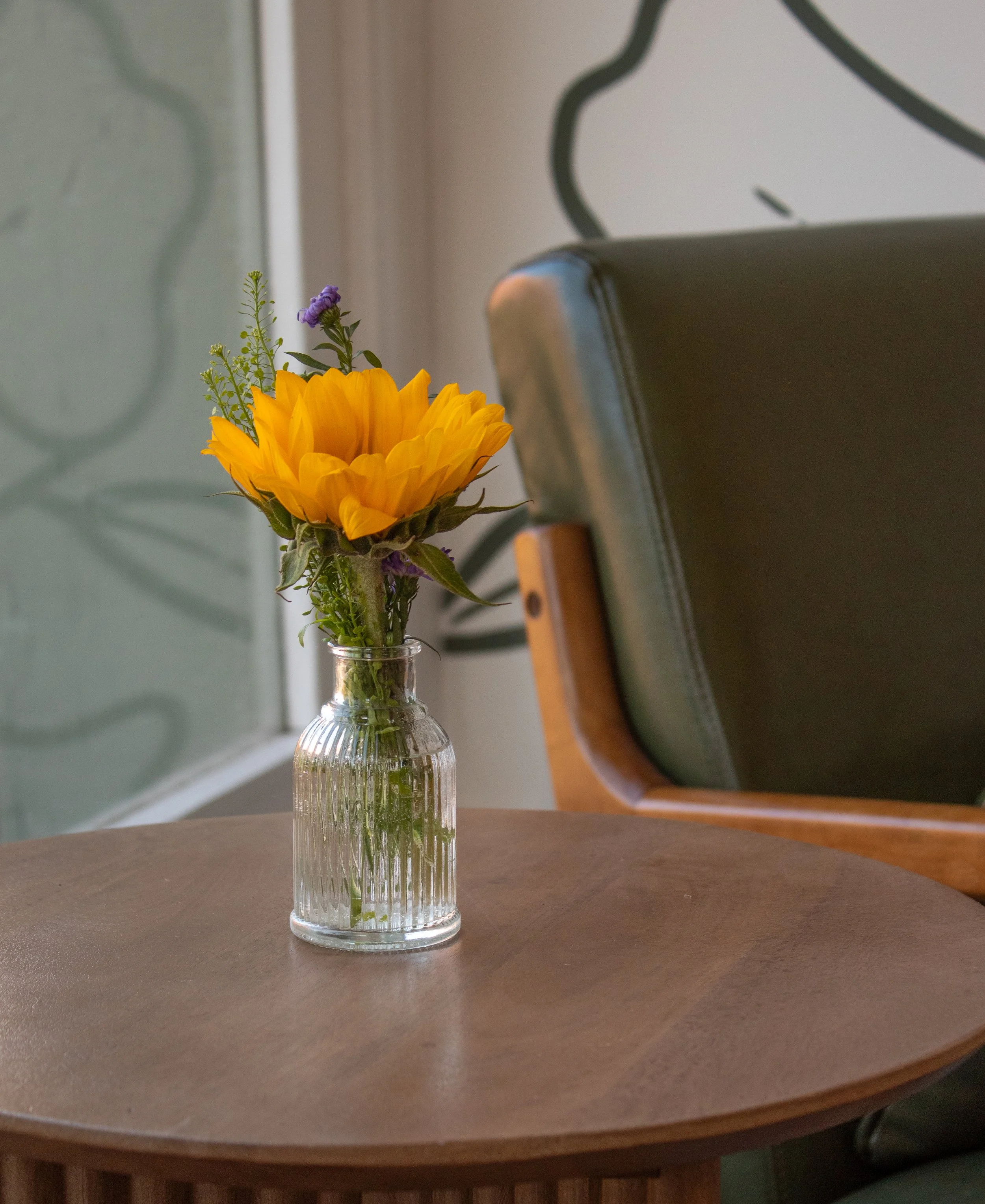 A small glass vase with a sunflower and purple flowers on a round wooden table.