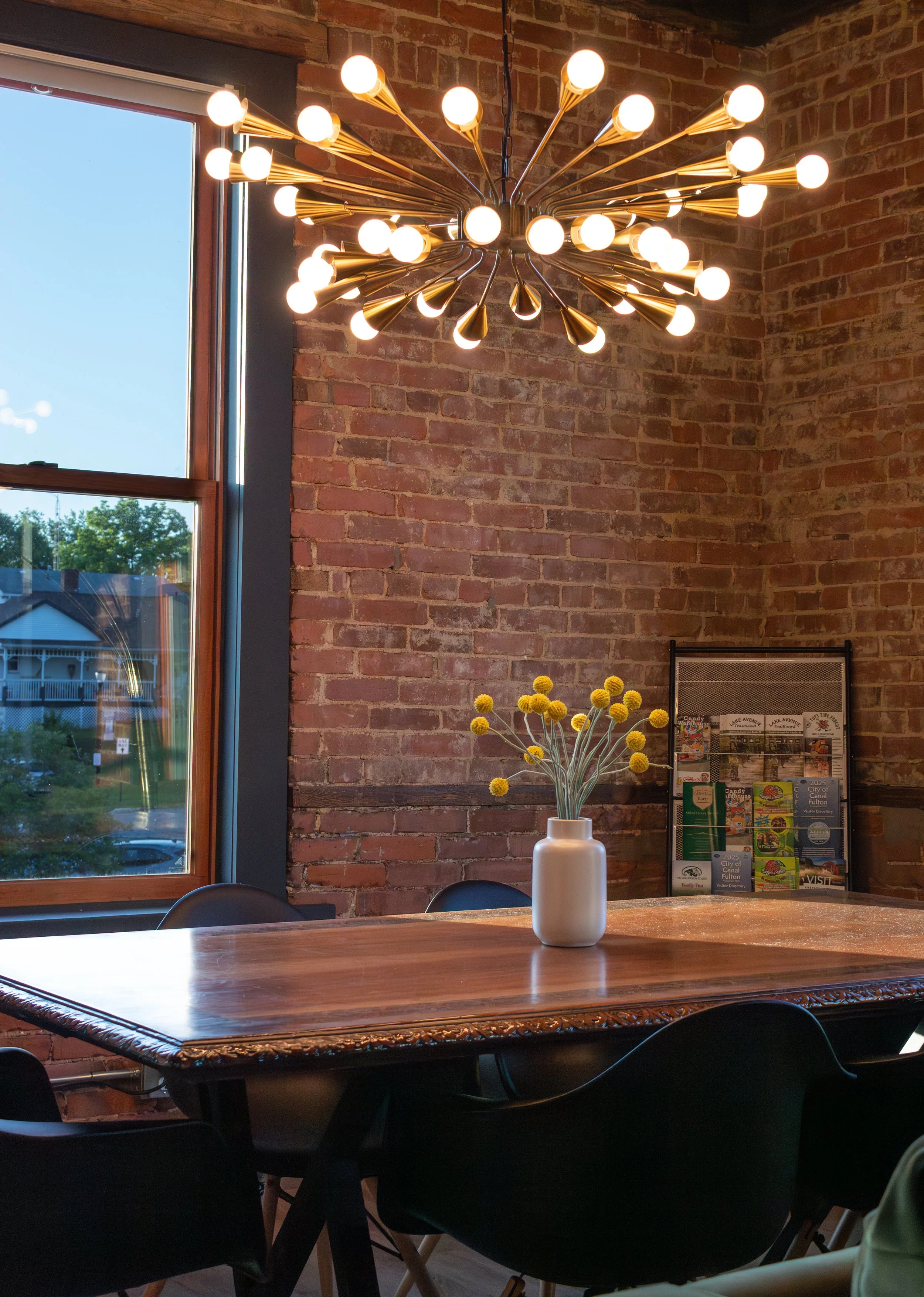 A modern interior dining area with a large wooden table, surrounded by black chairs, a white vase with yellow spherical flowers on the table, and a brick wall behind. A large window on the left shows a view of houses and trees outside. A unique chand