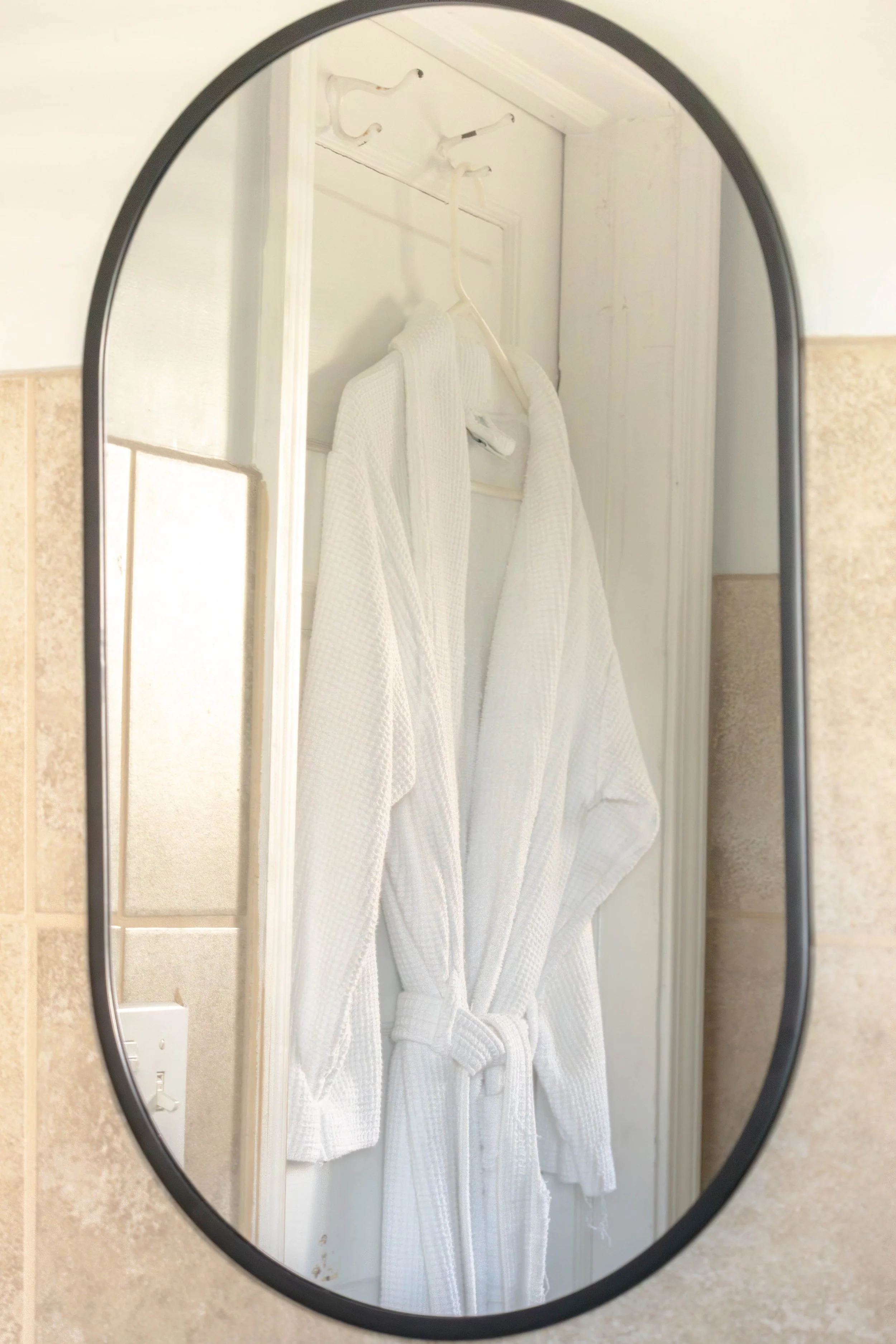 A bathroom mirror reflecting a white bathrobe hanging on a hook on a white door, with beige tiled walls surrounding the mirror.