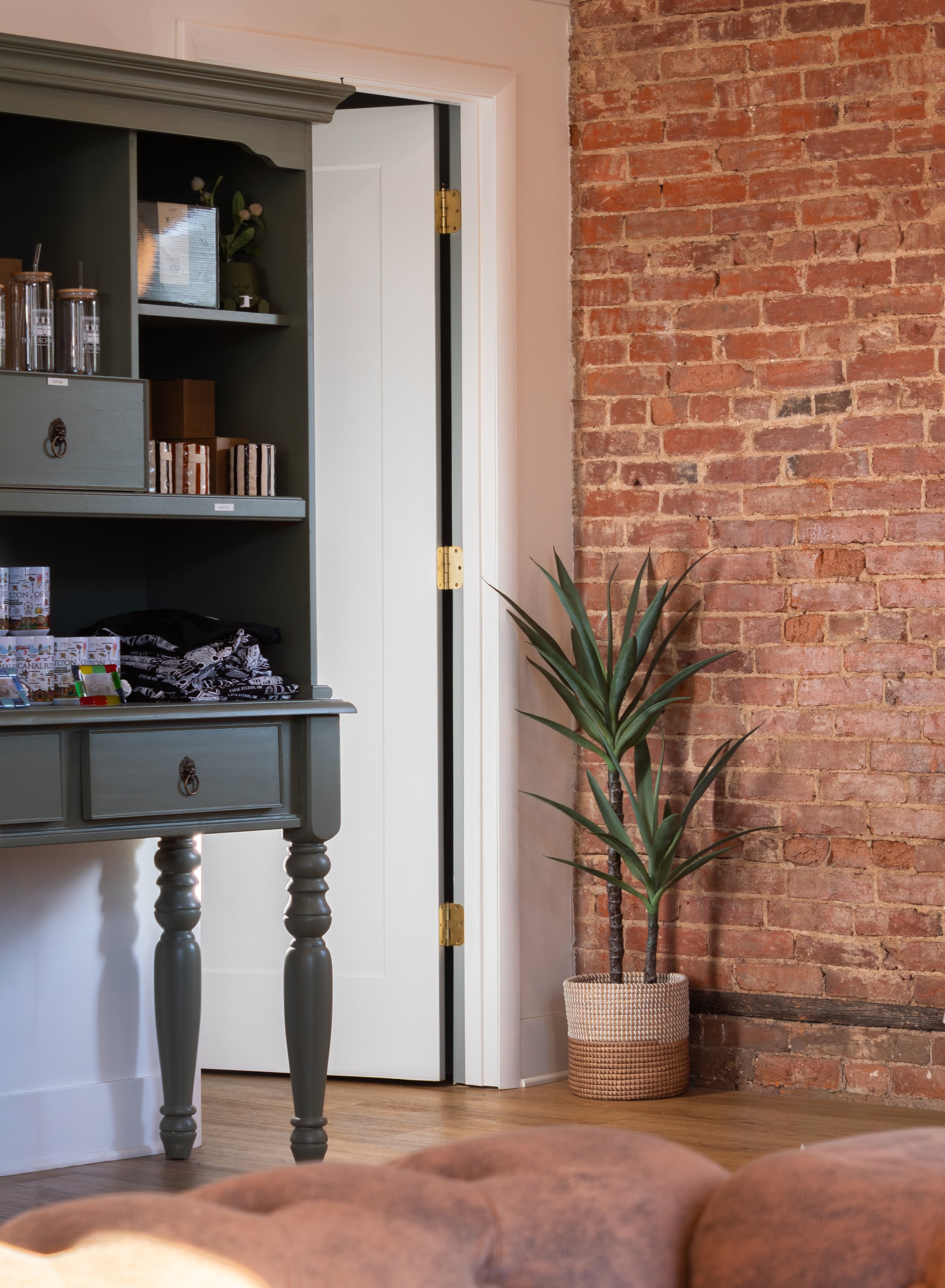 Interior view showing a brick wall on the right, a green wooden table with turned legs on the left, a white door in the background, a potted plant with long green leaves near the wall, and some decorative items on the table.