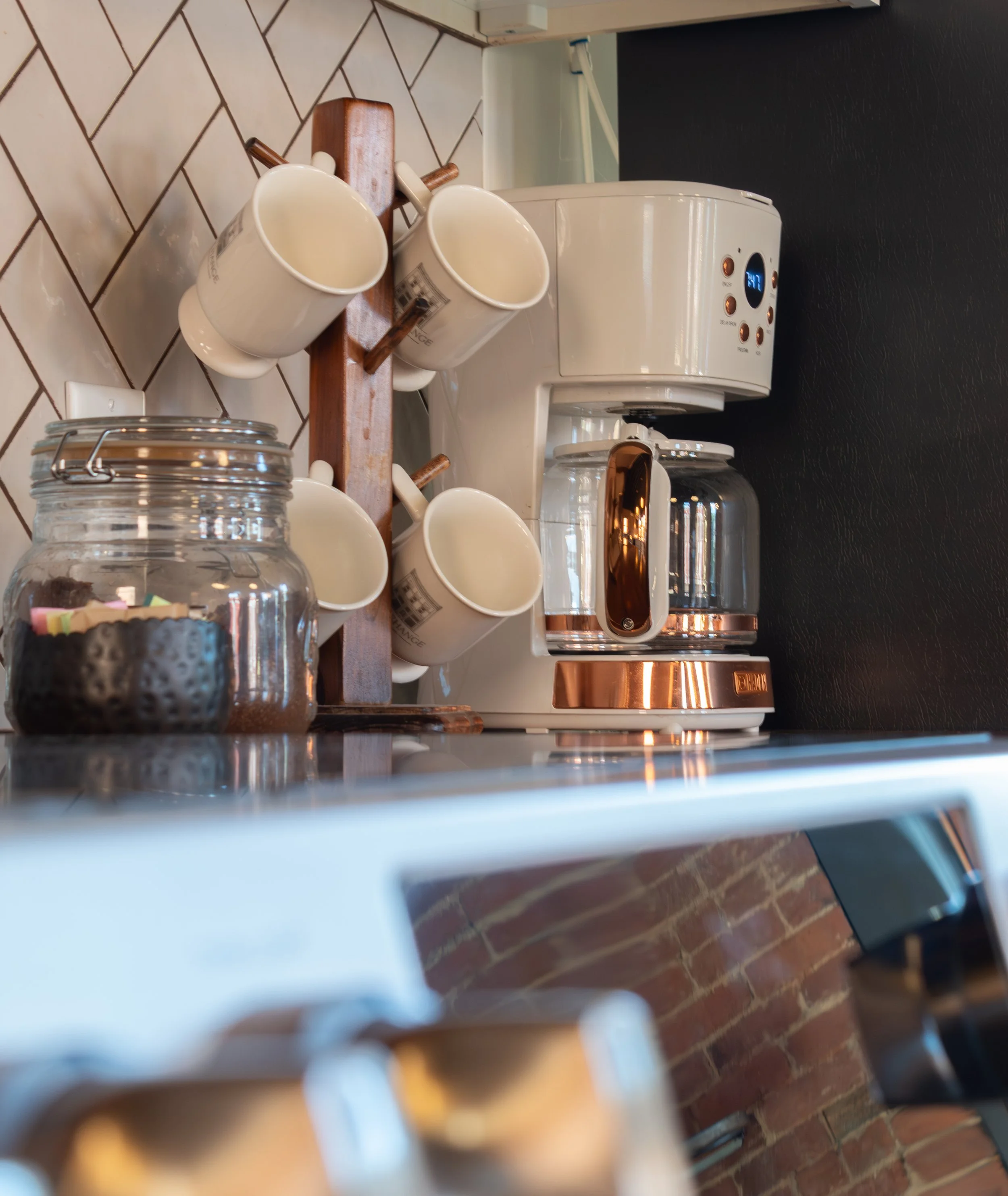 A row of white mugs with brown handles on a wooden tray next to a coffee machine on a kitchen countertop. The background shows a brick wall.