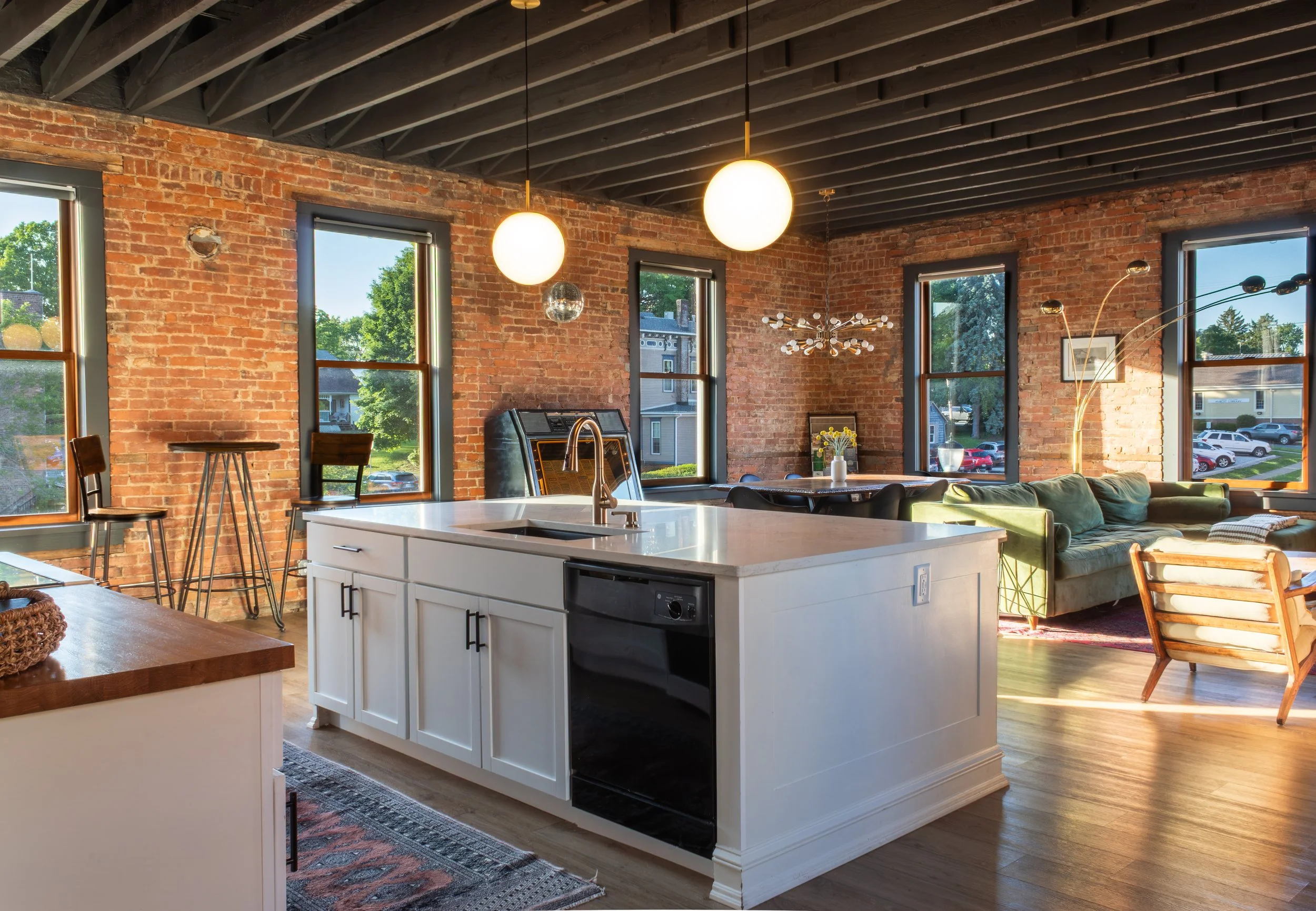 Open-concept kitchen and living area with exposed brick walls, large windows with views of trees and houses, pendant lights, and modern decor.
