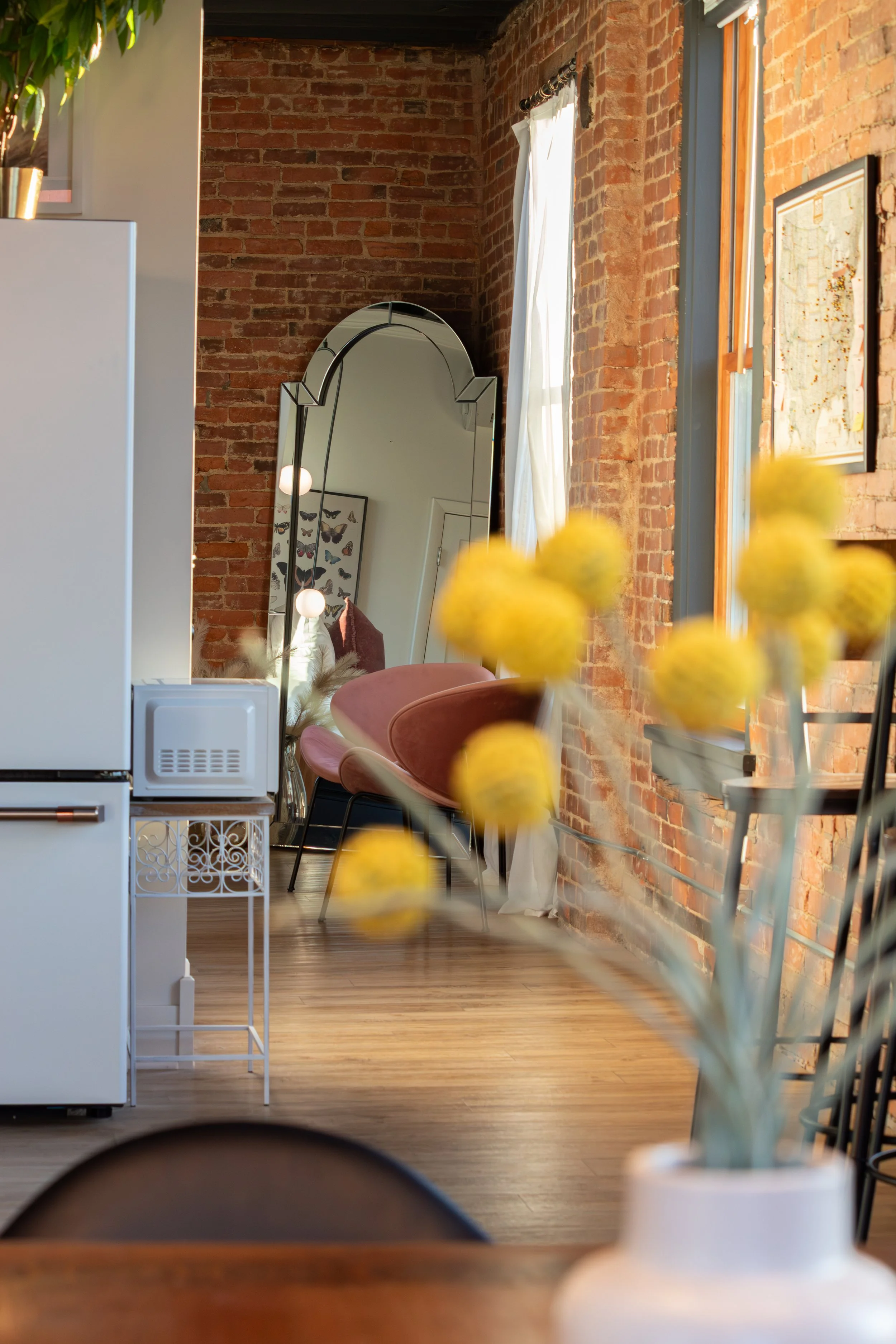 Interior of a room with exposed red brick walls, a large standing mirror, and a window with white curtains. Yellow flowers in a white vase are in the foreground.
