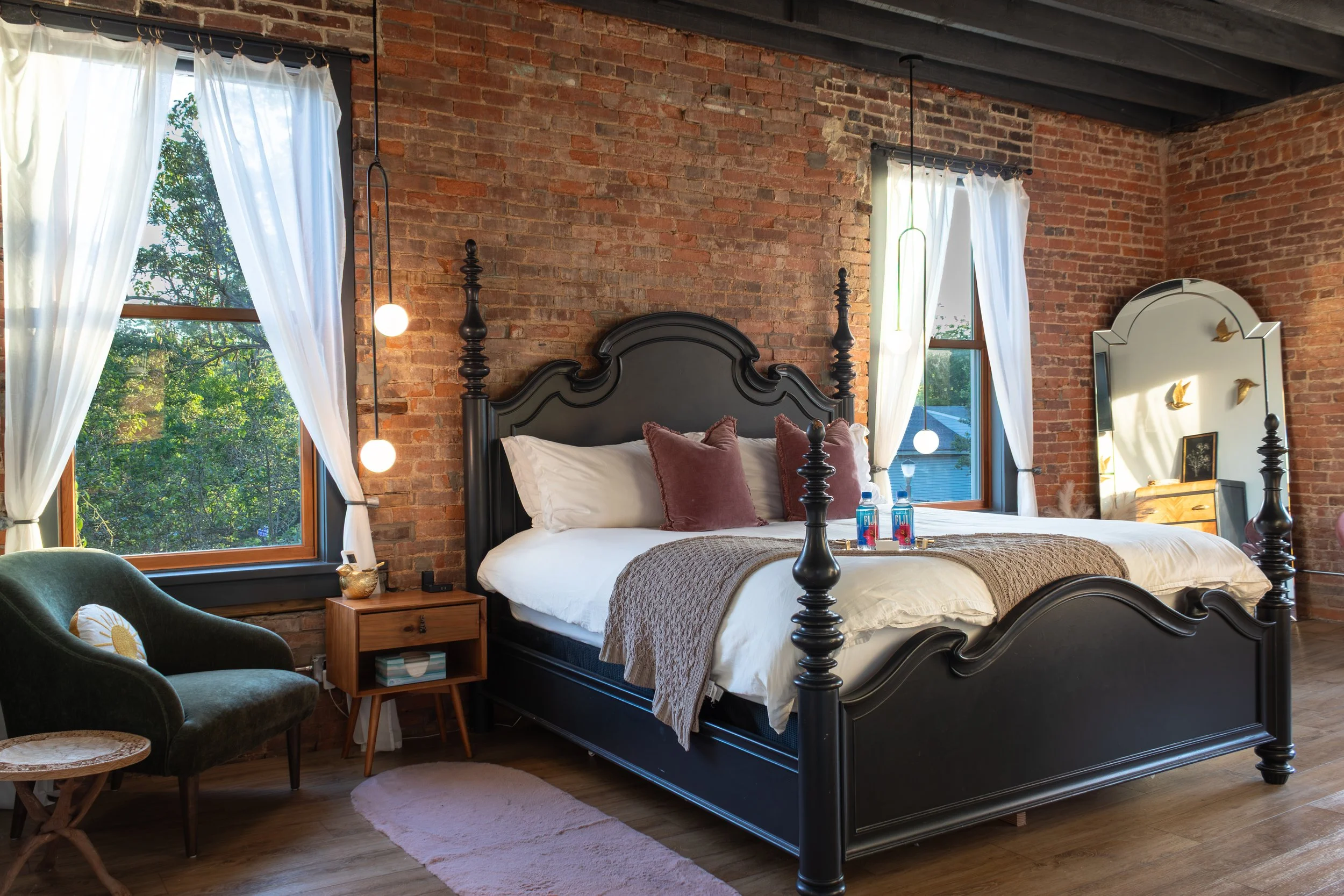 A bedroom with brick walls, a black wooden bed frame with white bedding and pink pillows, large windows with white curtains, a green upholstered chair, a small wooden side table, and a large mirror. There are two pendant lights hanging beside the bed