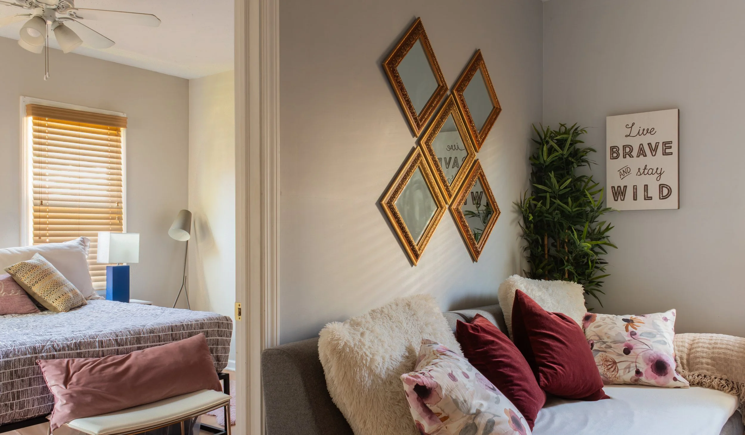 A cozy living space featuring a bed with pink and patterned pillows, beside a sitting area with a floral and velvet pillows. The room has a wall decor of four ornate, gold-framed mirrors, a white canvas with motivational quote, and a tall green plant