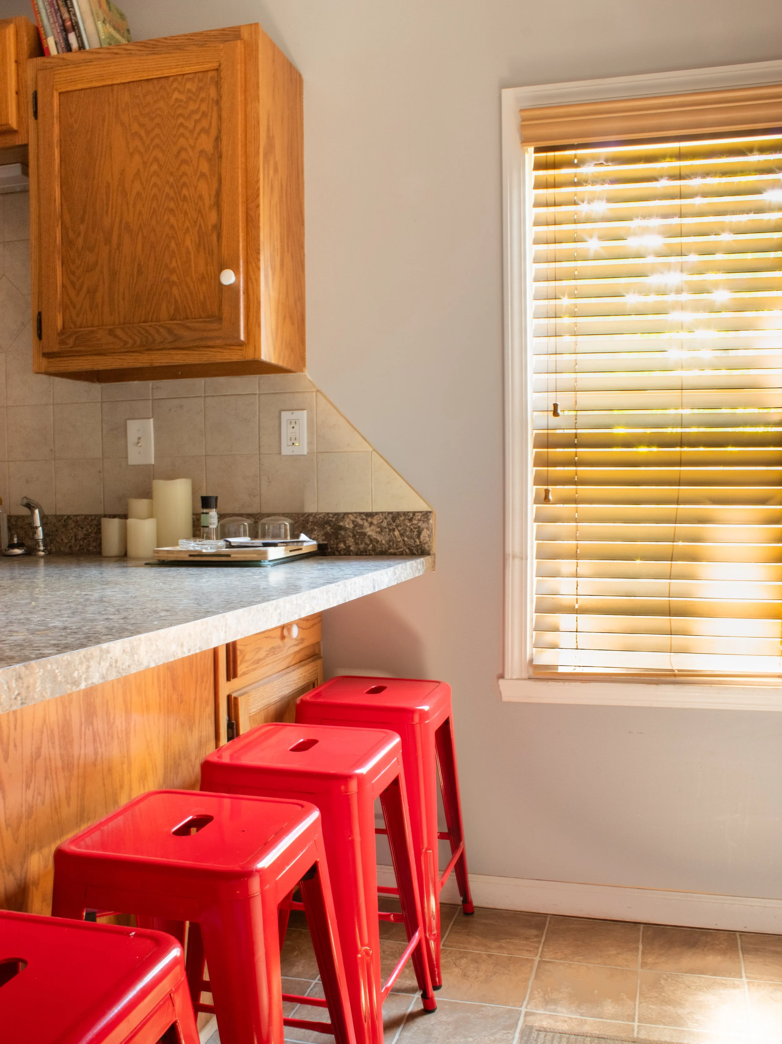 A kitchen corner with a granite countertop, wooden cabinets, candles, and two red stools in front. There's a window with partially closed blinds allowing sunlight to stream through.