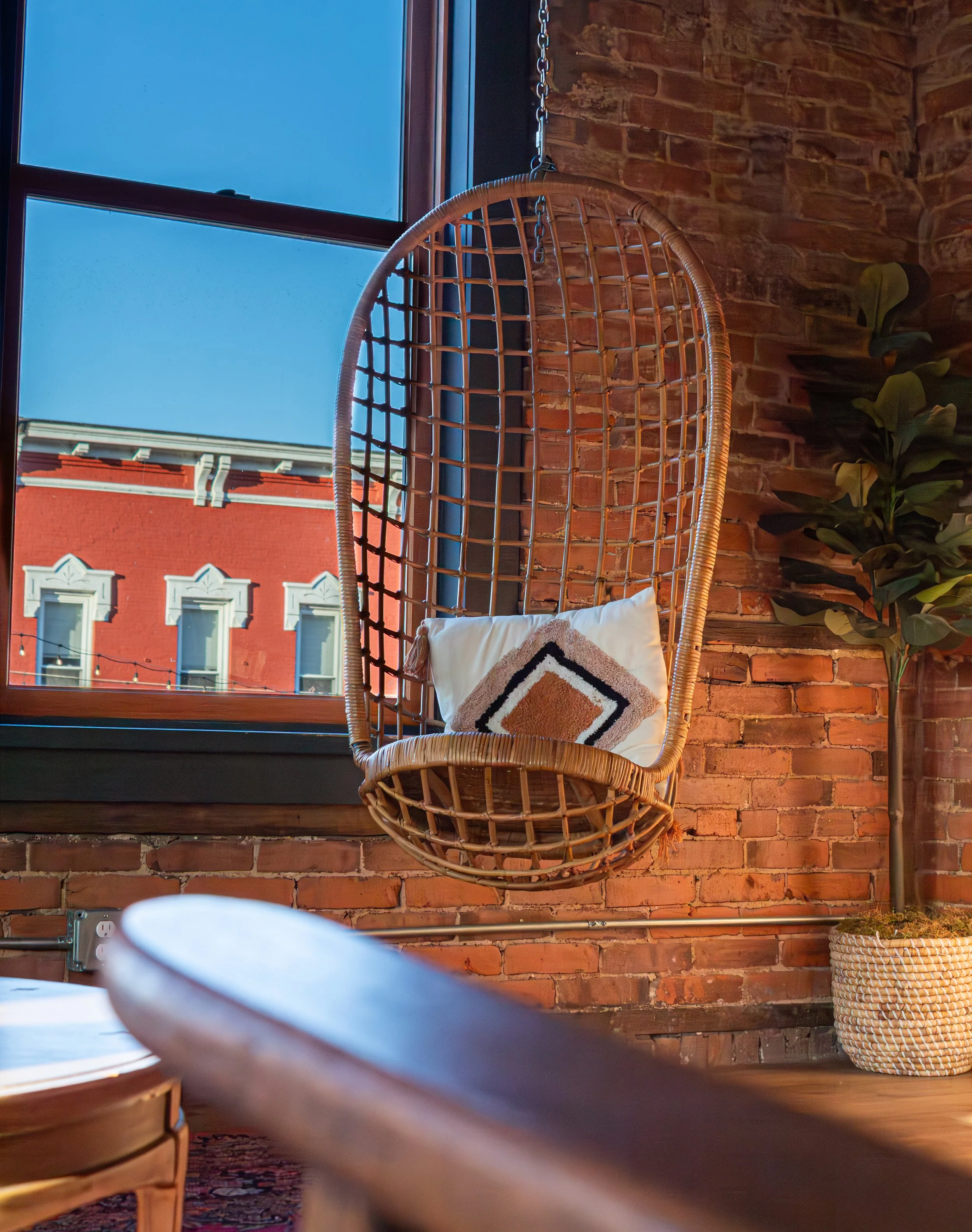 Indoor seating area with wicker chair, brick wall, window showing blue sky and part of red building exterior, plant to the right.