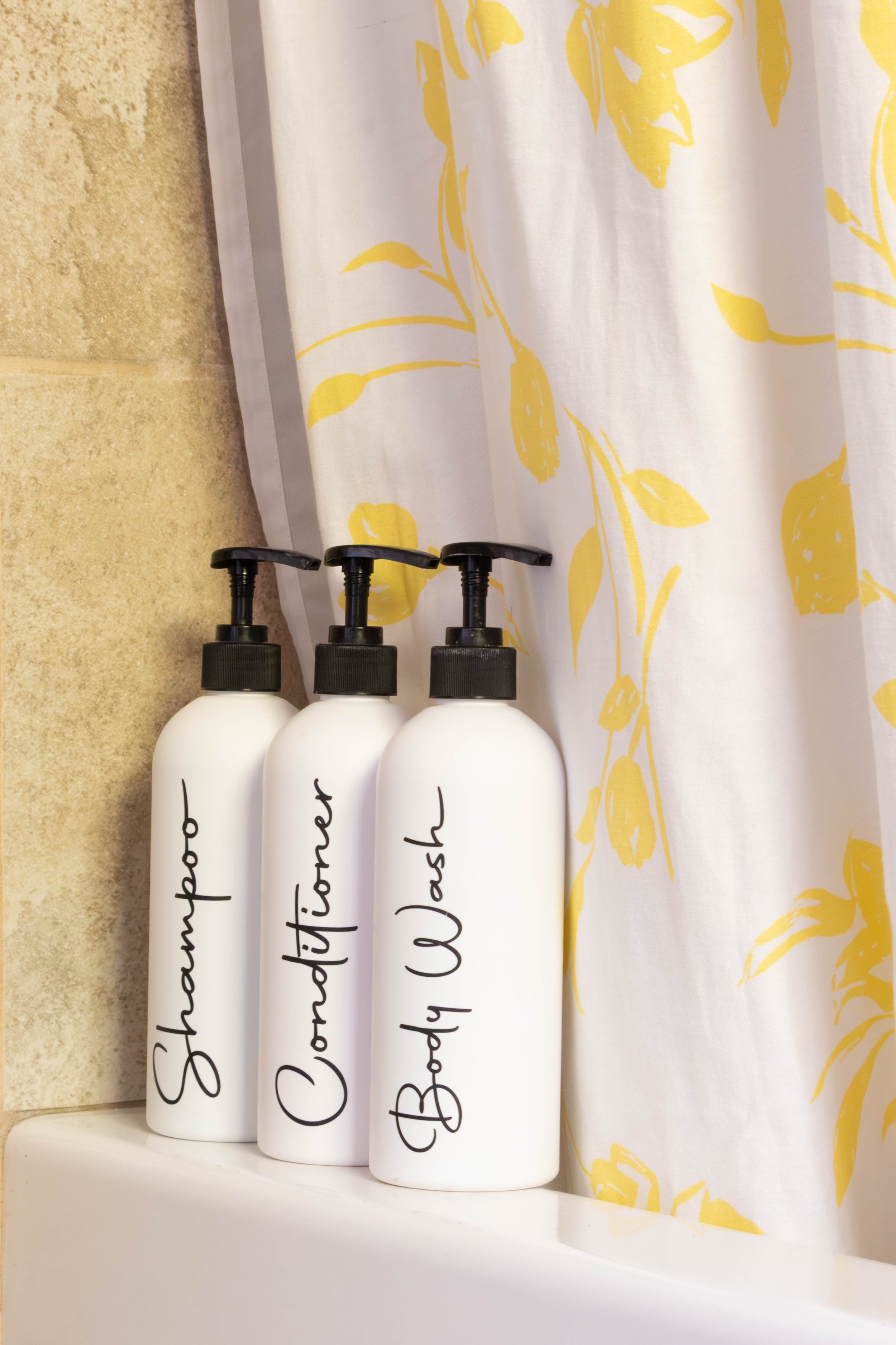 White bottles with black pump dispensers labeled with black cursive handwriting, placed on a white shelf near a white curtain with a yellow floral pattern.
