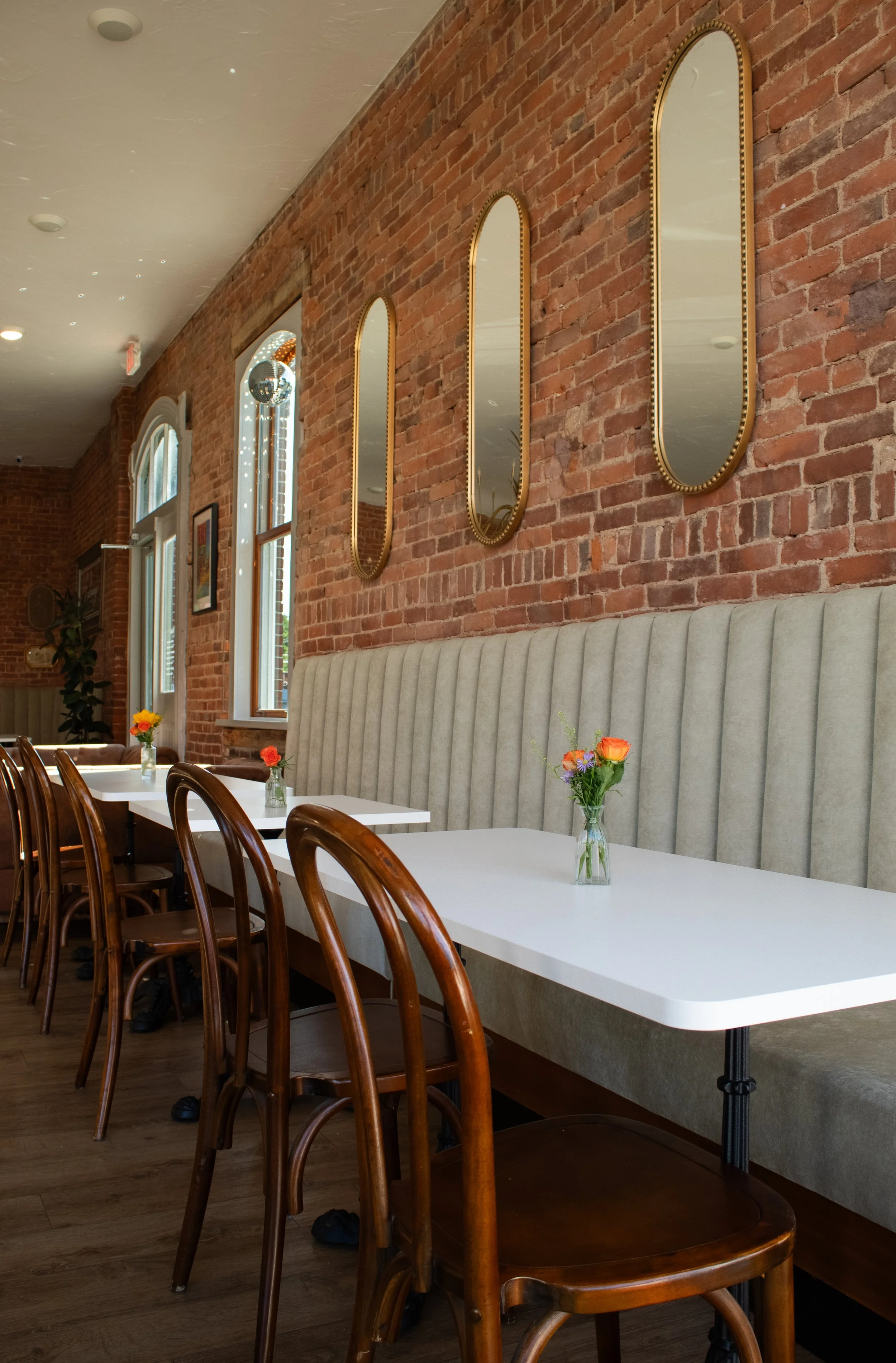 Interior of a restaurant or dining area with a long white table, small vases with flowers, wooden chairs, a beige upholstered bench, exposed brick wall with three decorative mirrors, large windows, and ceiling lights.