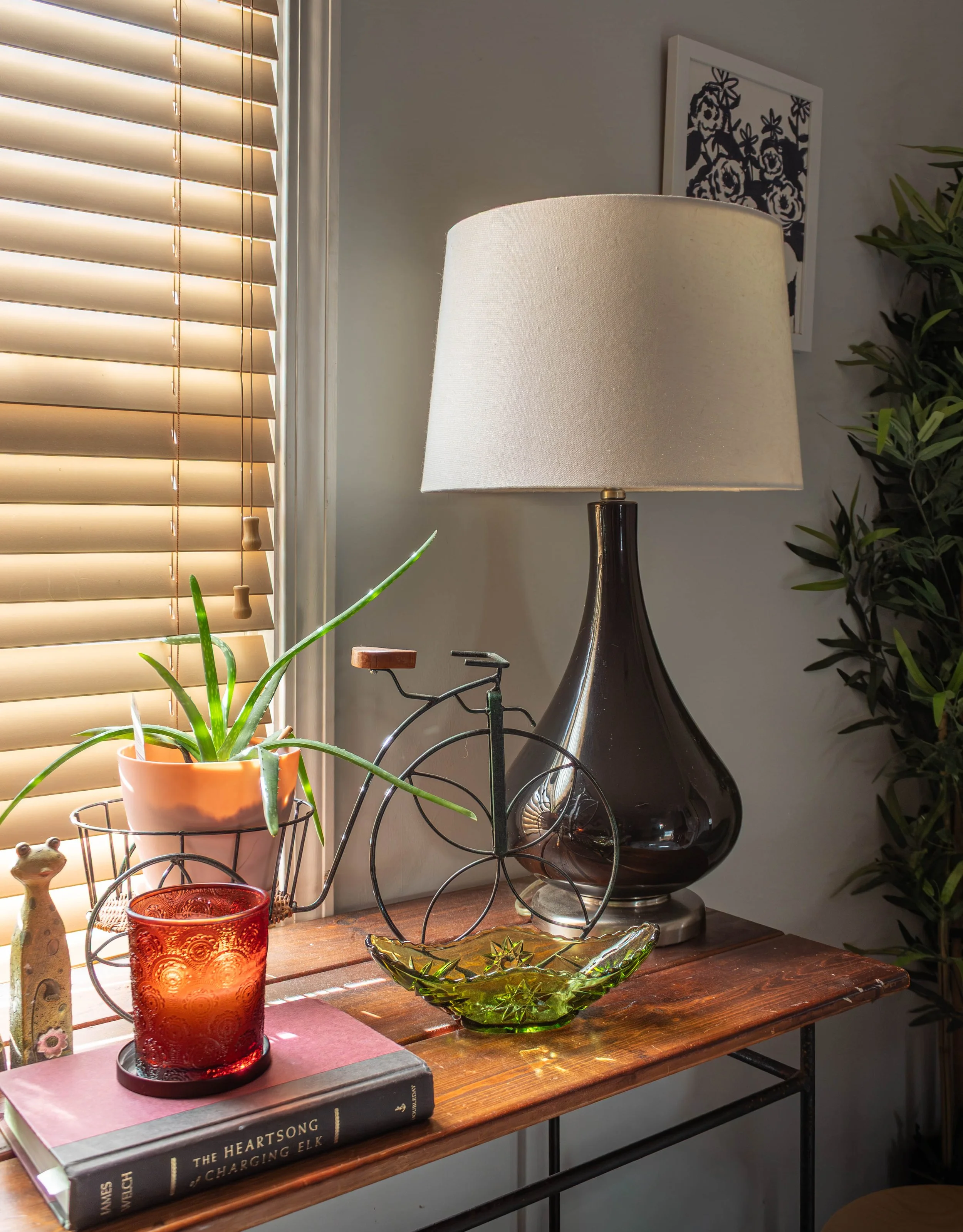 A wooden table holds decorative items, including a red glass candle holder, a green glass bowl, a potted aloe vera plant, and a small statue. Behind the table is a window with closed beige blinds. To the right, on a white wall, there is a framed blac