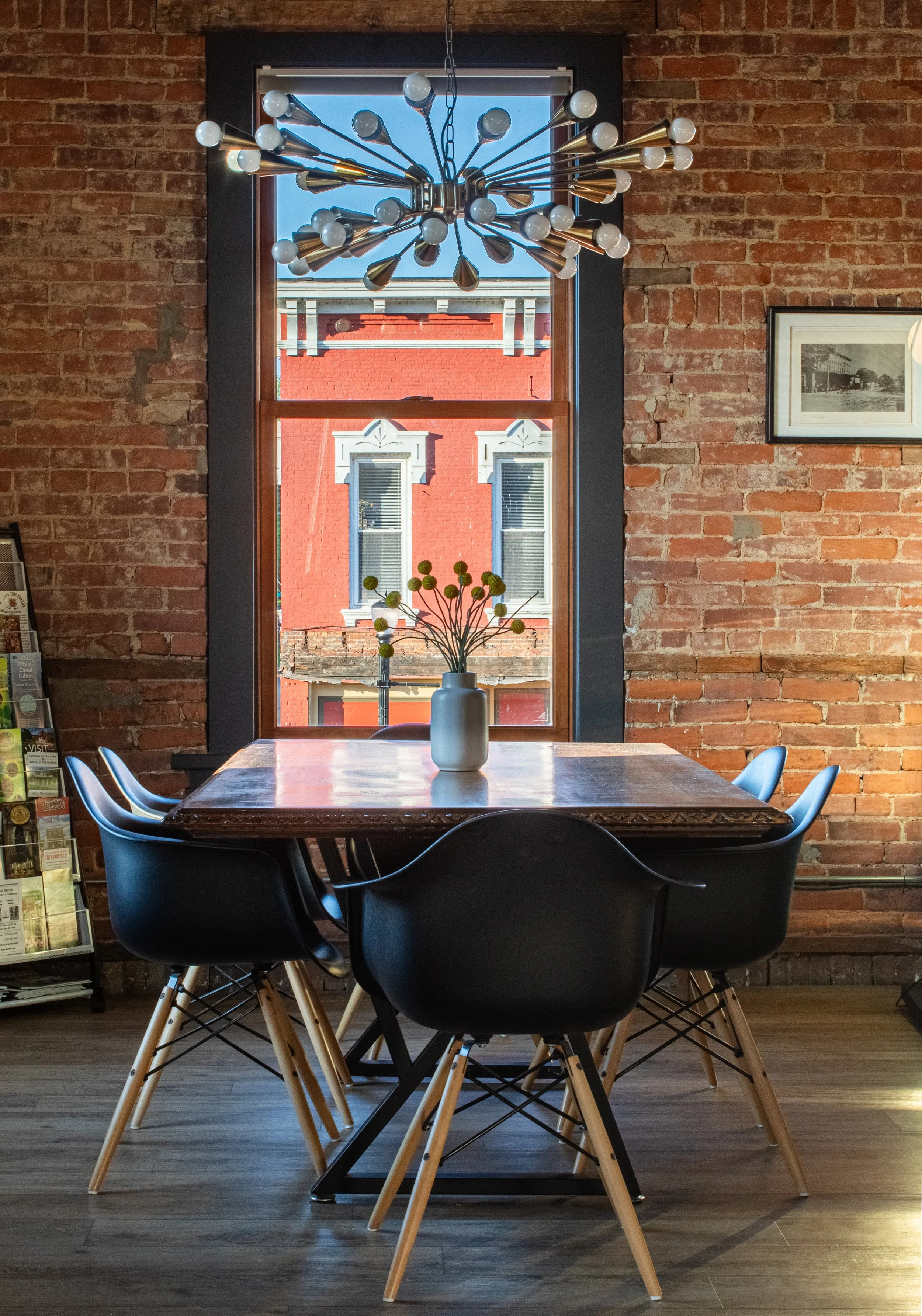 Interior of a restaurant with a table and six black chairs, a large window with a view of a red brick building, a modern chandelier hanging from the ceiling, a white vase with flowers on the table, and brick walls with a framed black-and-white photo.