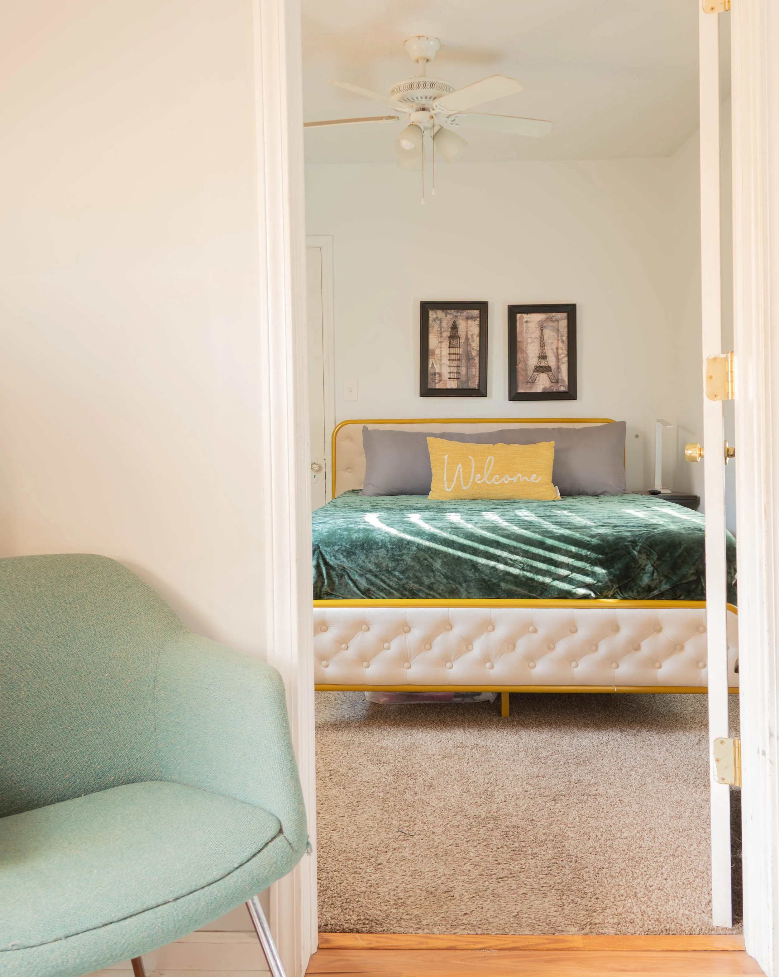 A bedroom with a bed, wall art, and a ceiling fan, viewed through an open door with a green chair in the foreground.