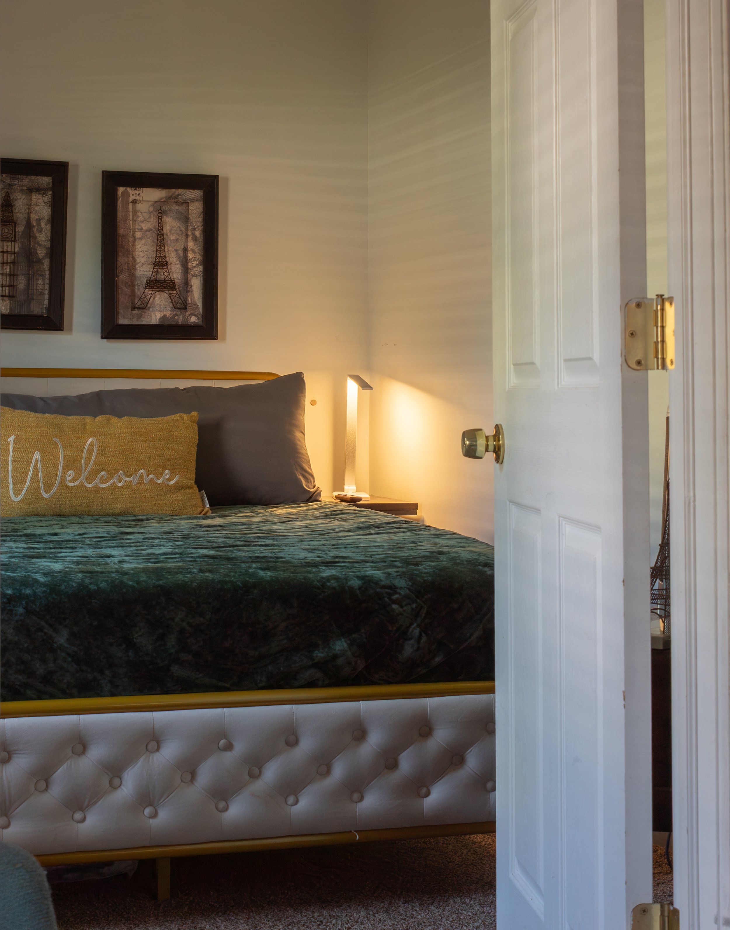 A bedroom with a white tufted bed frame, beige carpet, light-colored wall with framed artwork, bedside table with a lamp, and an open white door.