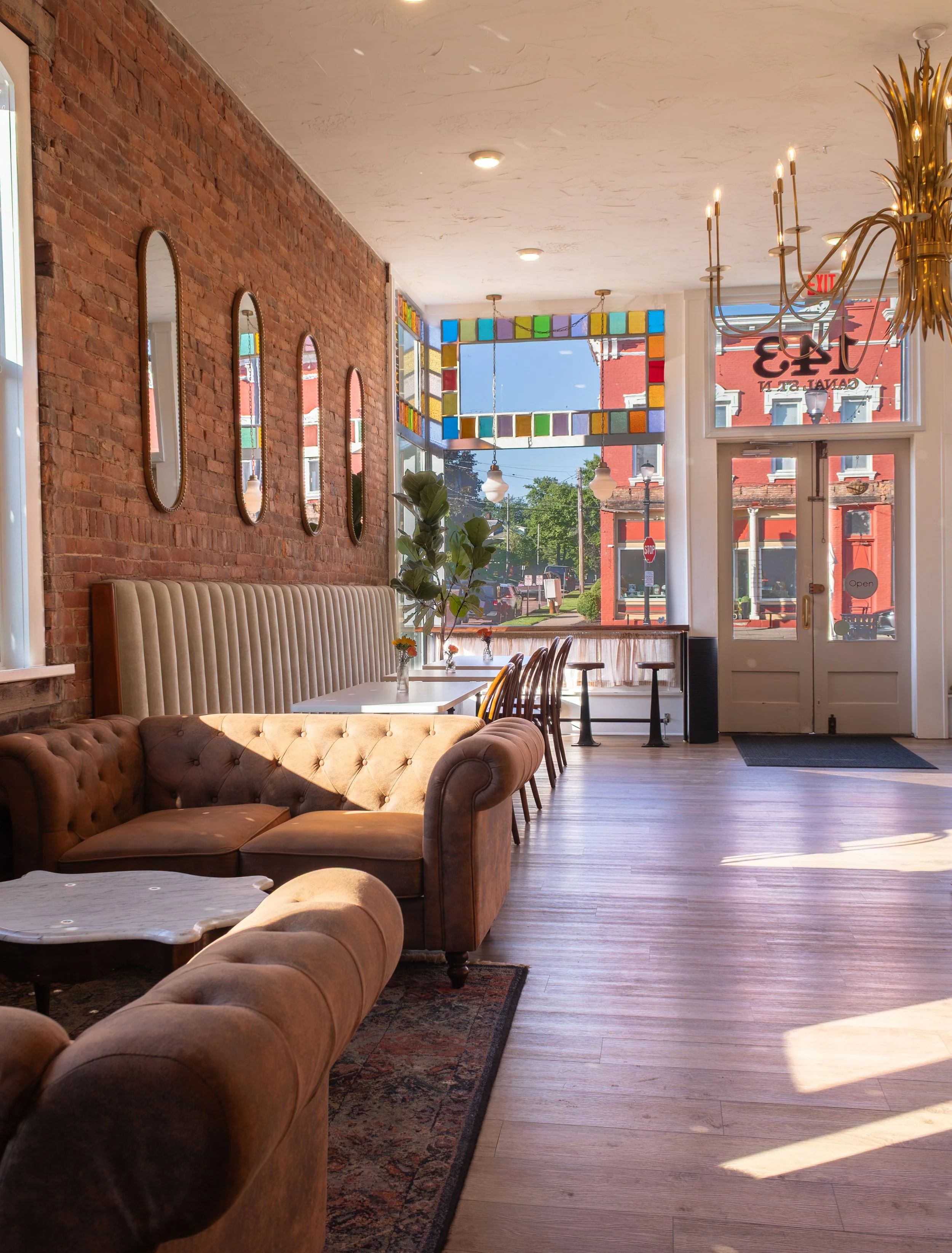 Interior of a café with a brick wall on one side, decorated with vertical mirrors. There are tables with vases of flowers, and a leather sofa. Large windows let in natural light, showing a city street outside. A stained glass fixture and chandeliers 