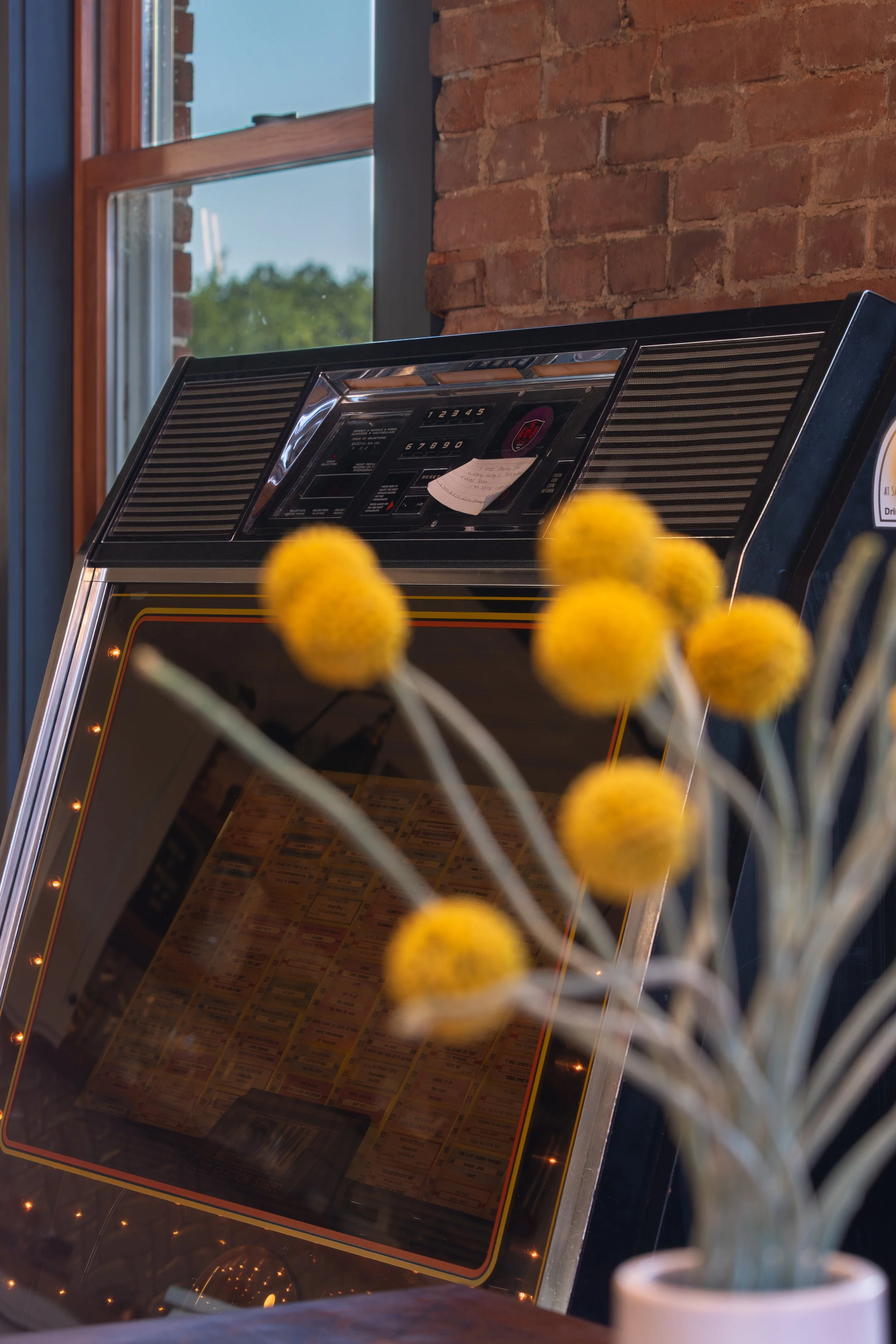 A vintage jukebox with a black top and chrome accents, partially obscured by yellow flowers in a white vase, set against a brick wall and a window with a view of greenery outside.