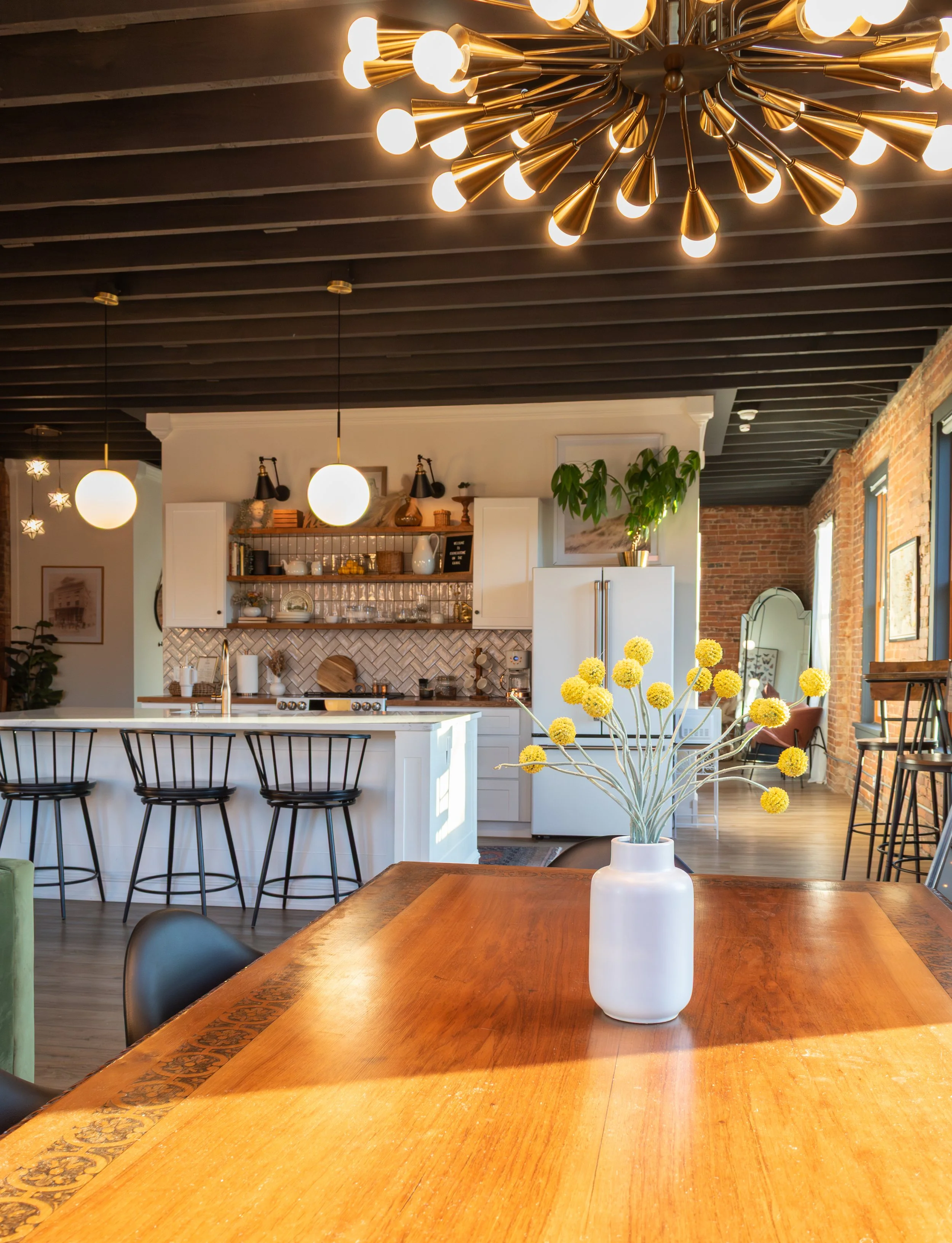 A bright kitchen with modern light fixtures, a white kitchen island, white cabinets, yellow flowers in a white vase on a wooden table, and brick walls.