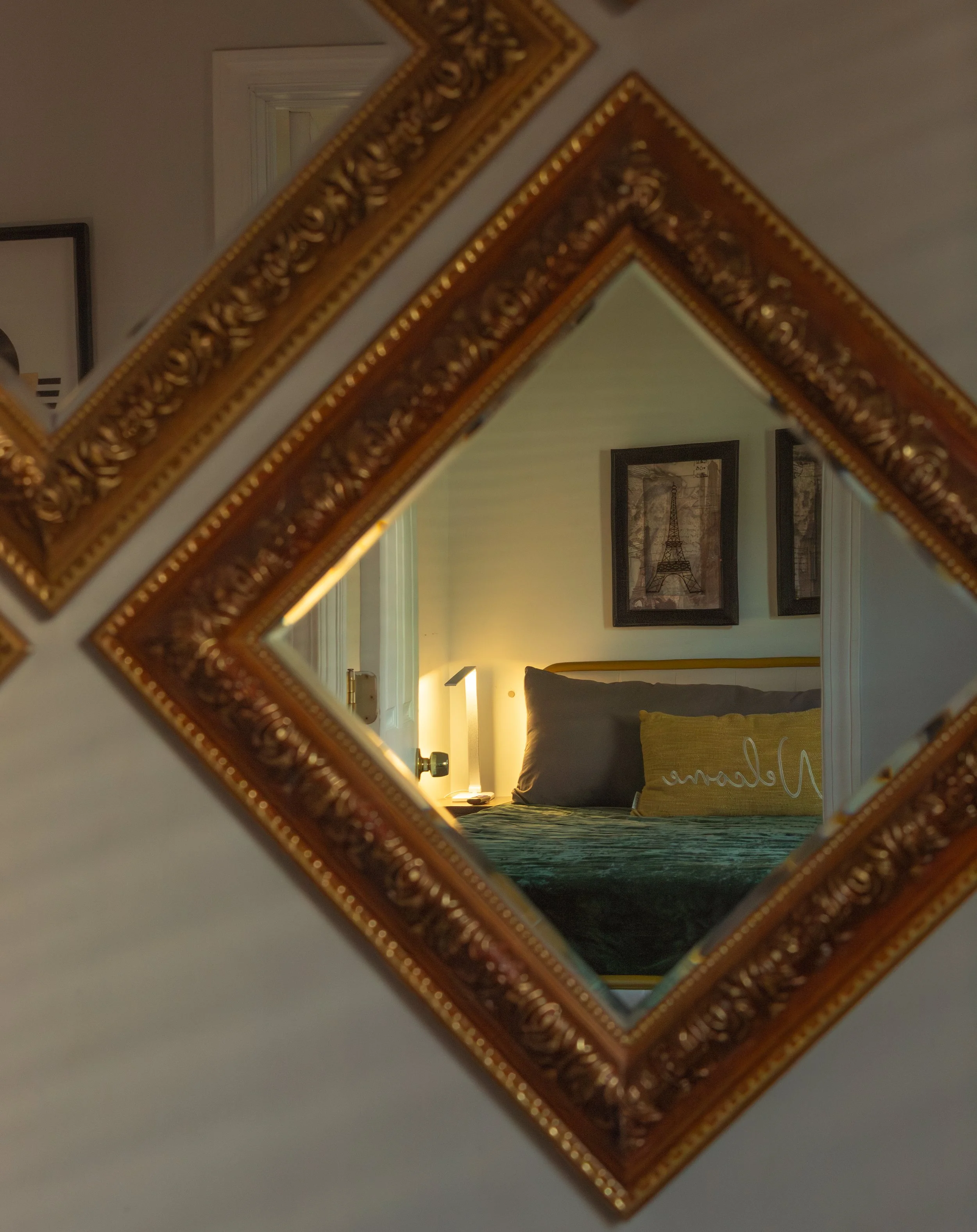 A reflection of a bedroom seen through a decorative mirror with a gold ornate frame. The room has framed pictures on the wall, a bed with pillows, and a bedside lamp.