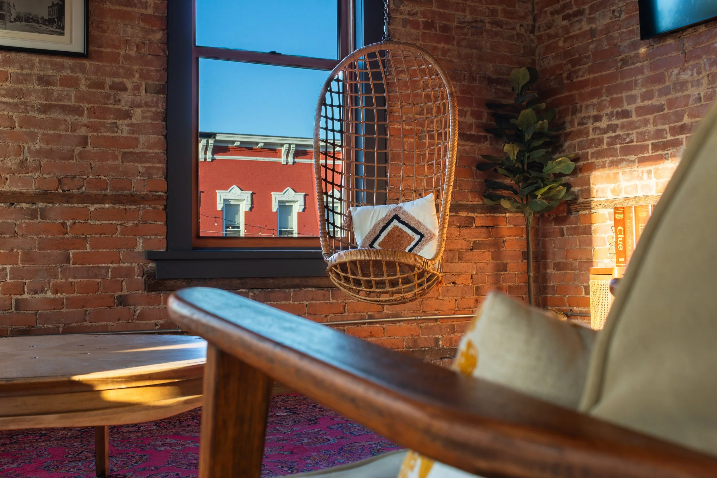 Interior of a room with brick walls, a window showing neighboring buildings, a rattan hanging chair with a cushion, a plant, and a bookshelf with orange books.