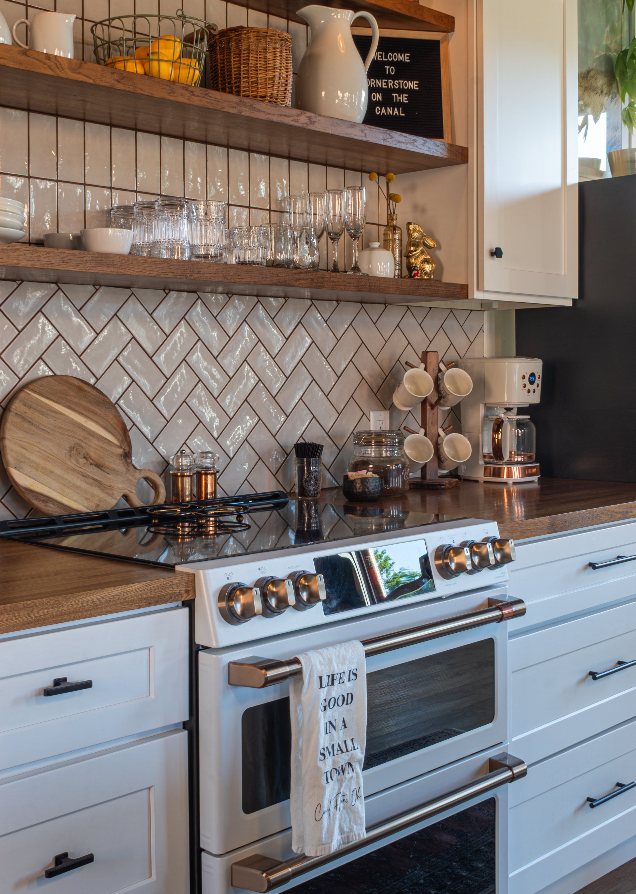 Kitchen with white stove, wooden countertops, and open shelves holding glasses, bowls, mugs, and a pitcher. There are white tiled walls and decorative items including a small blackboard with writing, a wicker basket, a ceramic pitcher, and a golden r