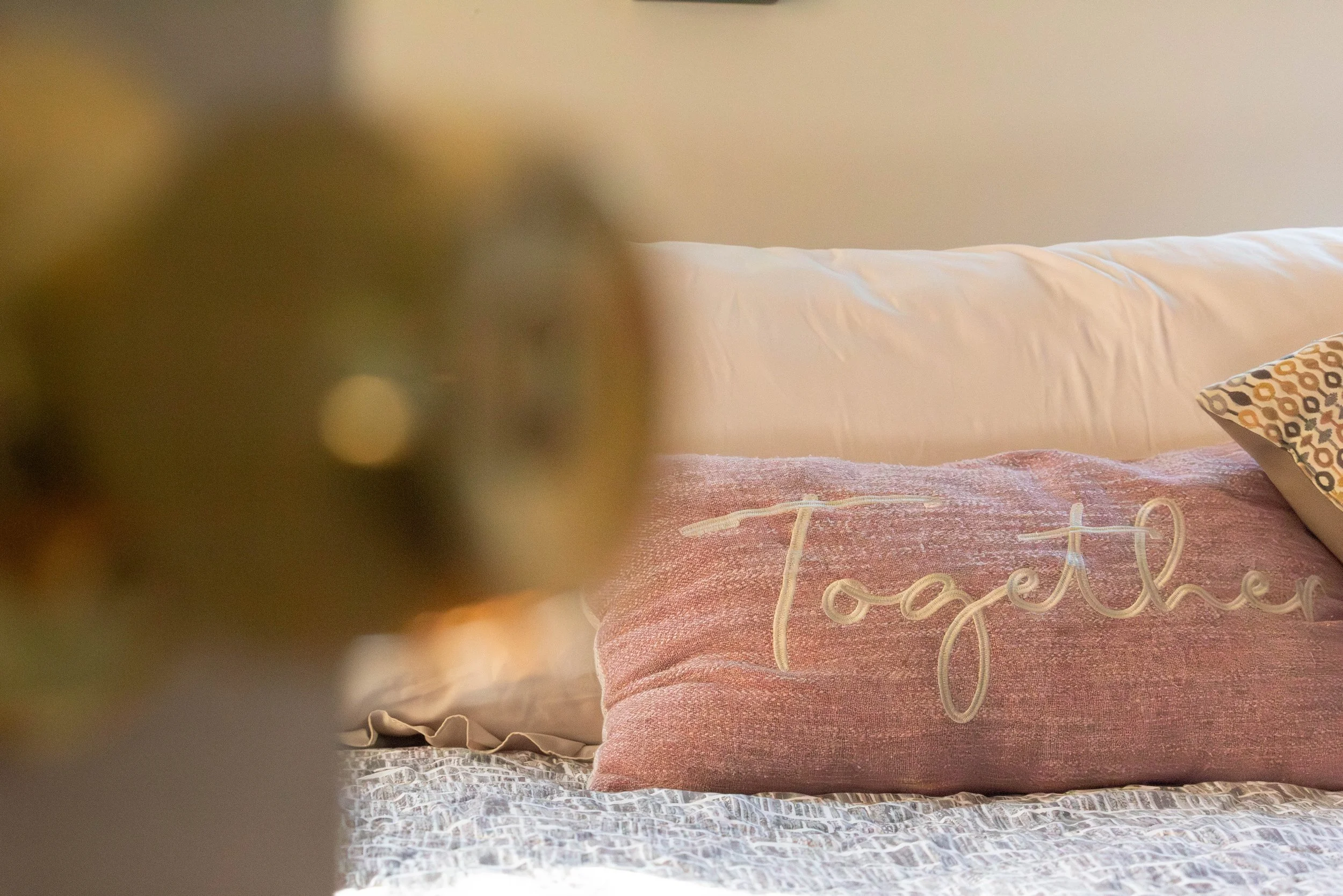 Close-up of a bed with a pink fabric comforter that has the word "Love" embroidered on it, white sheets, and a decorative pillow with a geometric pattern resting against the headboard. In the background, there is a beige headboard, a section of a wal