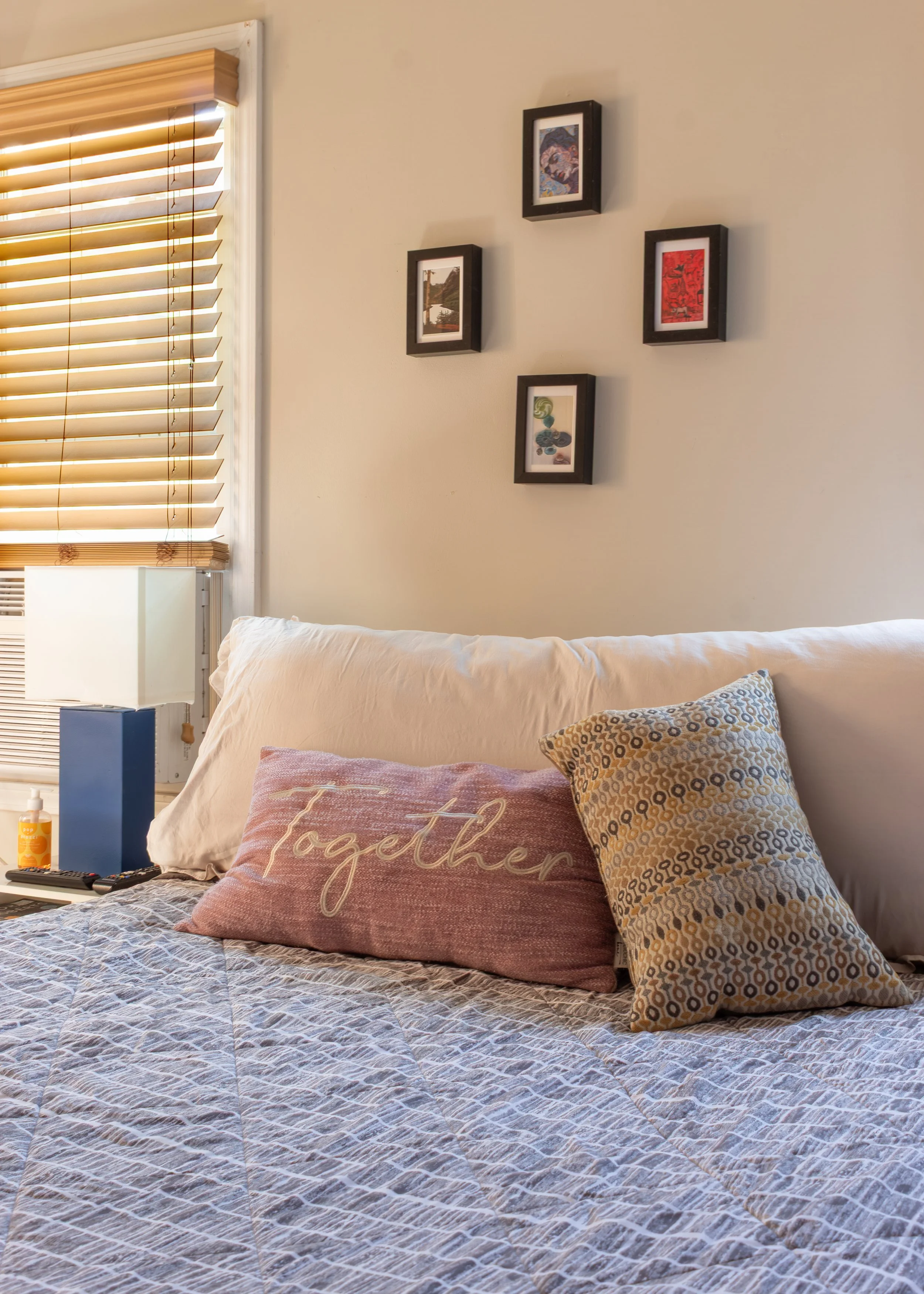 A bedroom with a bed that has decorative pillows including one with the words "together" and another with a patterned design. There are framed pictures on the wall and a window with wooden blinds letting in sunlight.