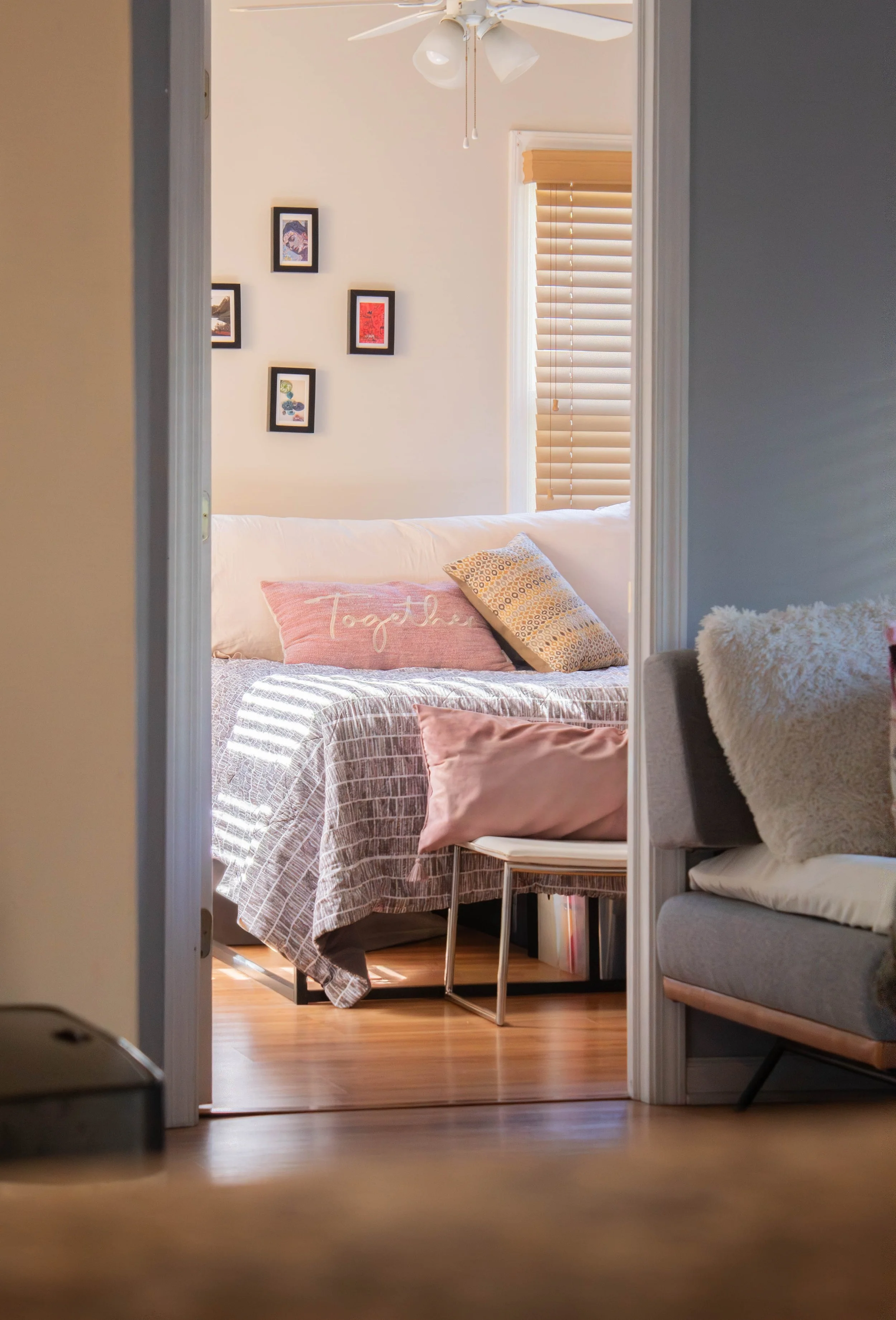A view into a cozy living room with a bed or couch with pillows, including a pink pillow with the word 'together' embroidered on it, a small side table, and a painting hanging on a wall with a ceiling fan overhead. There are wooden blinds on a window